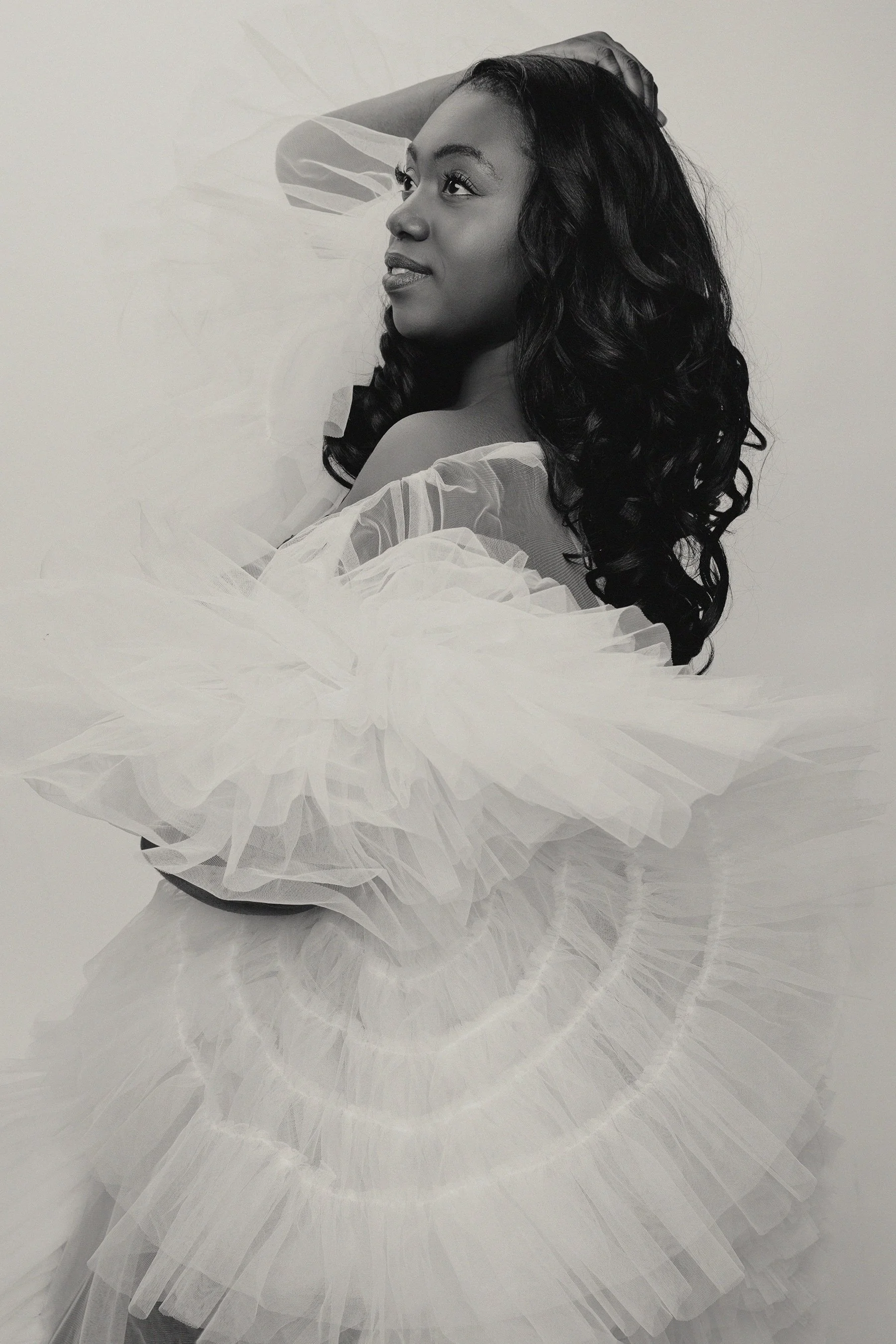 Black woman with long curly hair standing sideways wearing a dress made of multiple layers of tulle fabric.
