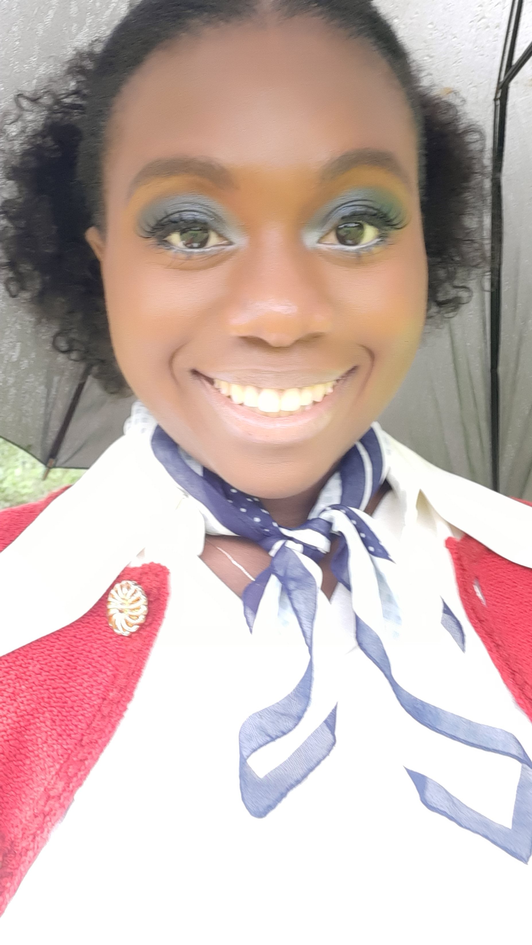 Close-up of a woman smiling with colorful makeup, wearing a white shirt, red blazer, and a blue and white neck scarf, holding an umbrella.