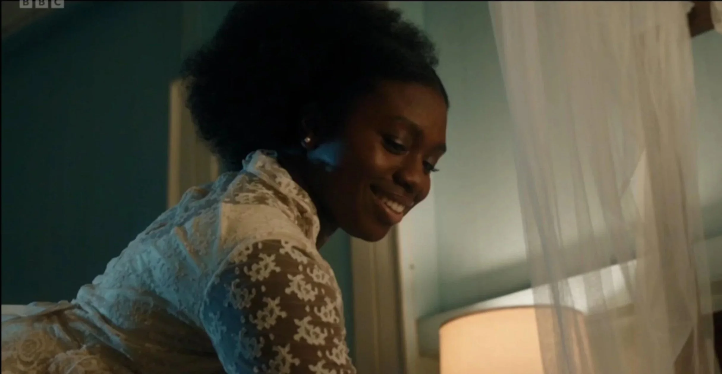 A woman with dark skin and natural hair smiling while sitting in a room, with a green wall, a beige curtains, and a lamp in the background.