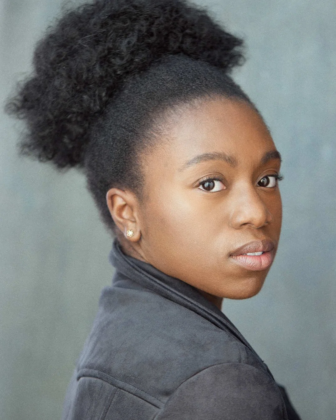 A young woman with natural curly hair styled in an updo, wearing a dark jacket and small earrings, looking over her shoulder at the camera.
