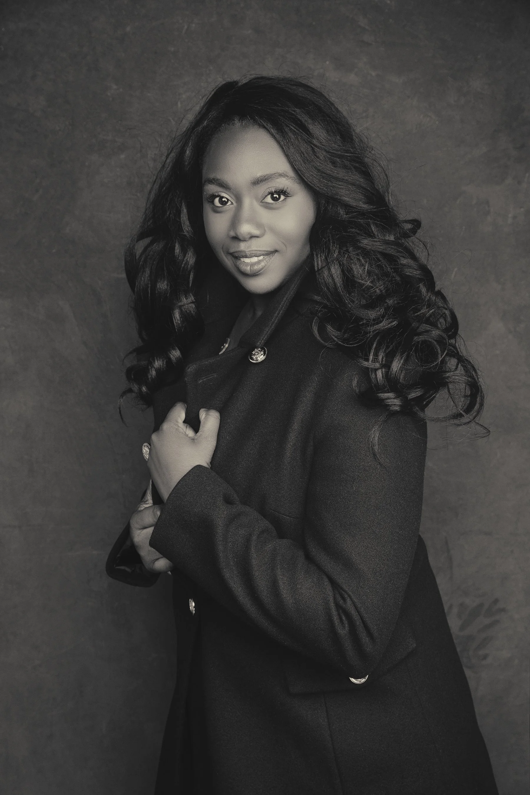 Black and white portrait of a woman with long curly hair, wearing a dark coat, smiling gently, against a plain dark background.