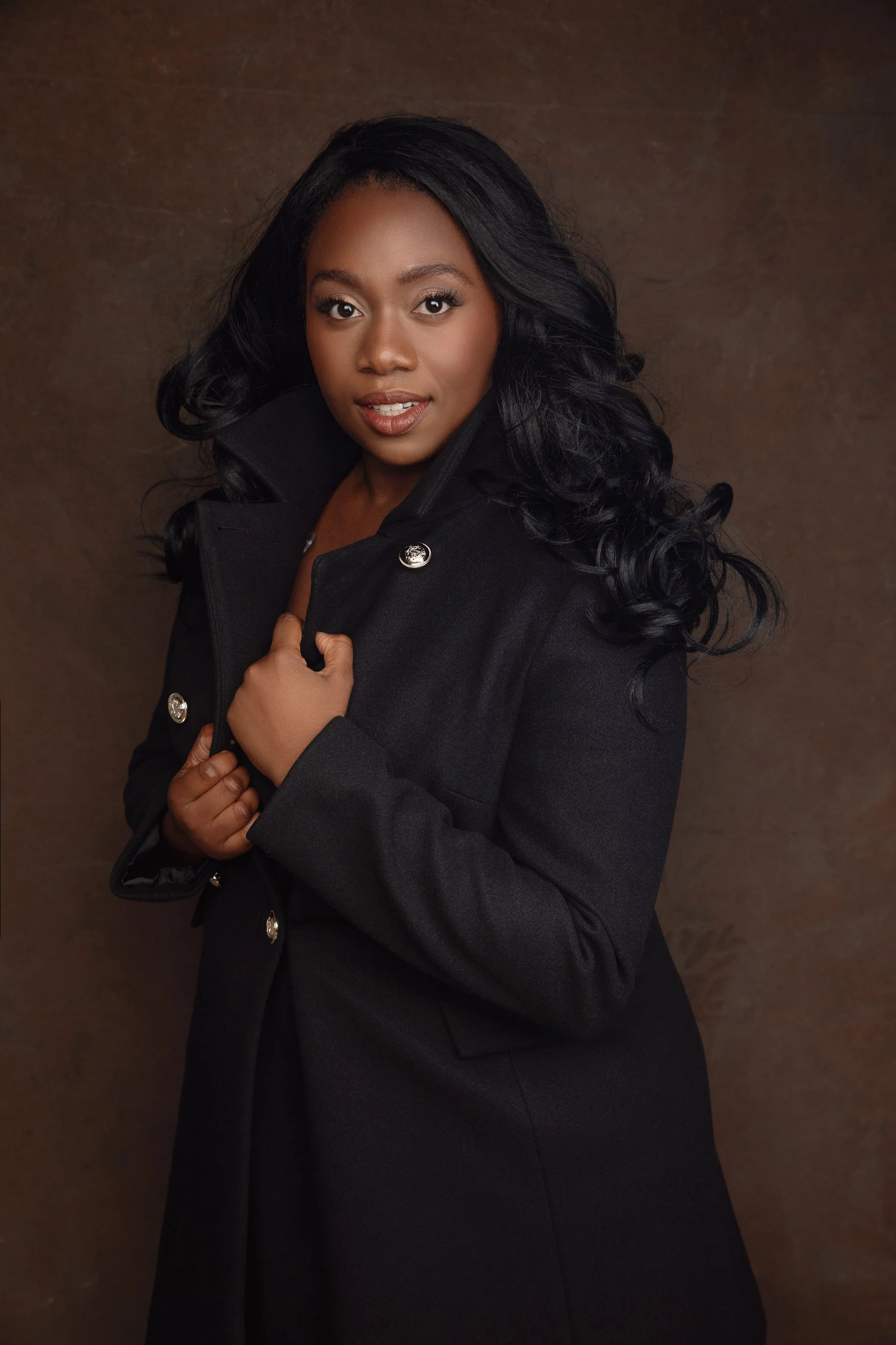 Portrait of a woman with long black wavy hair wearing a black coat with metallic buttons, posing against a brown textured background.