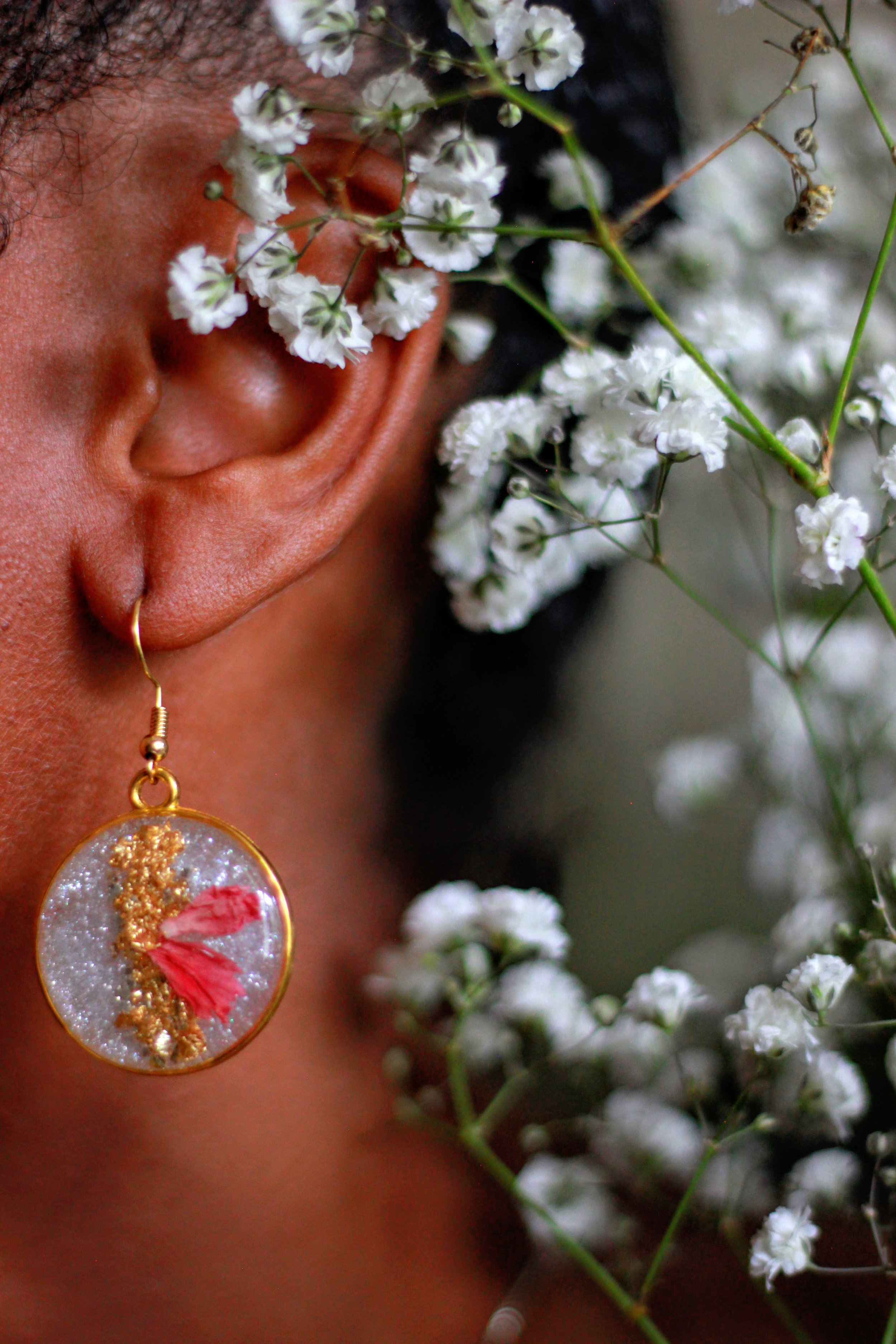 A person with dark skin and curly hair is wearing a gold hoop earring and a round pendant necklace. They are holding a bunch of white baby's breath flowers close to their face, partially obscuring it.