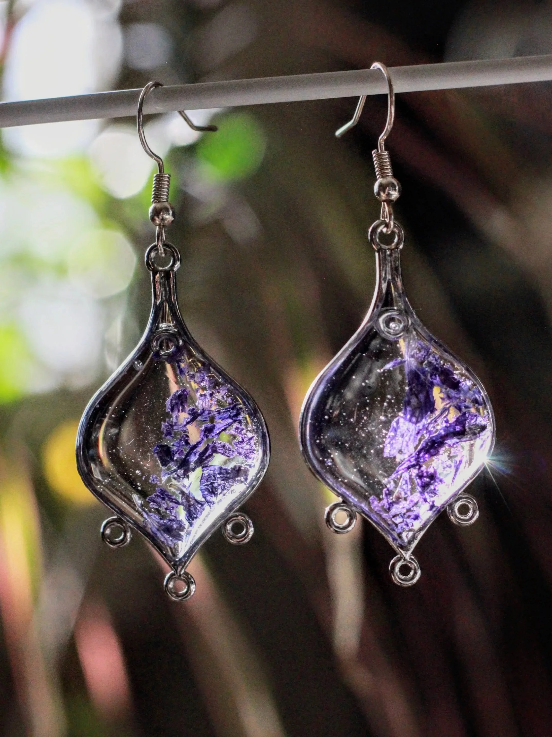 Close-up of silver drop-shaped earrings with purple and gold leaf patterns hanging from a thin metal rod.