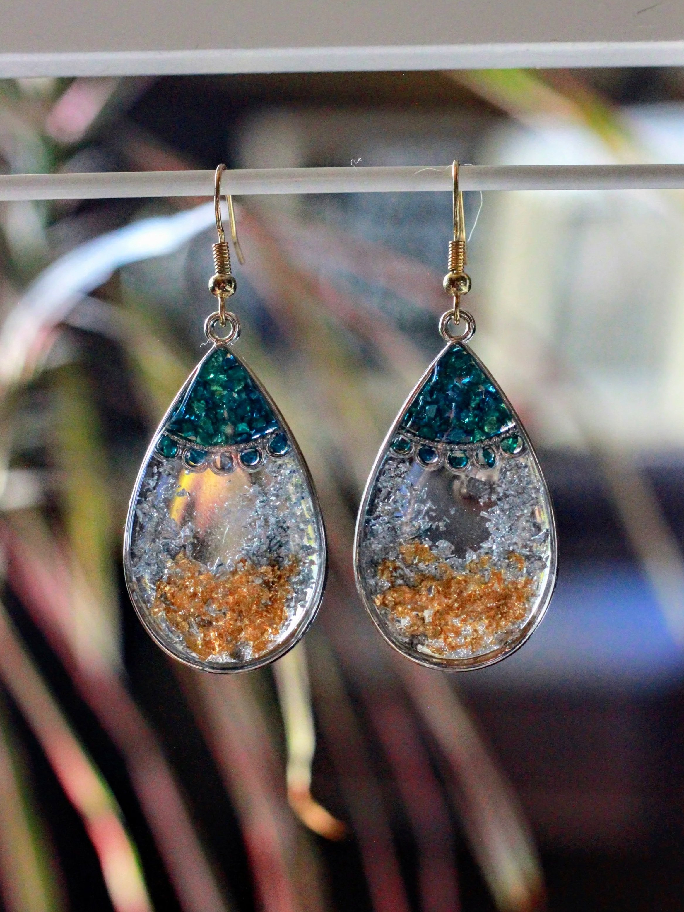 A pair of teardrop-shaped earrings with multicolored resin and embedded metallic flakes hanging from hooks.
