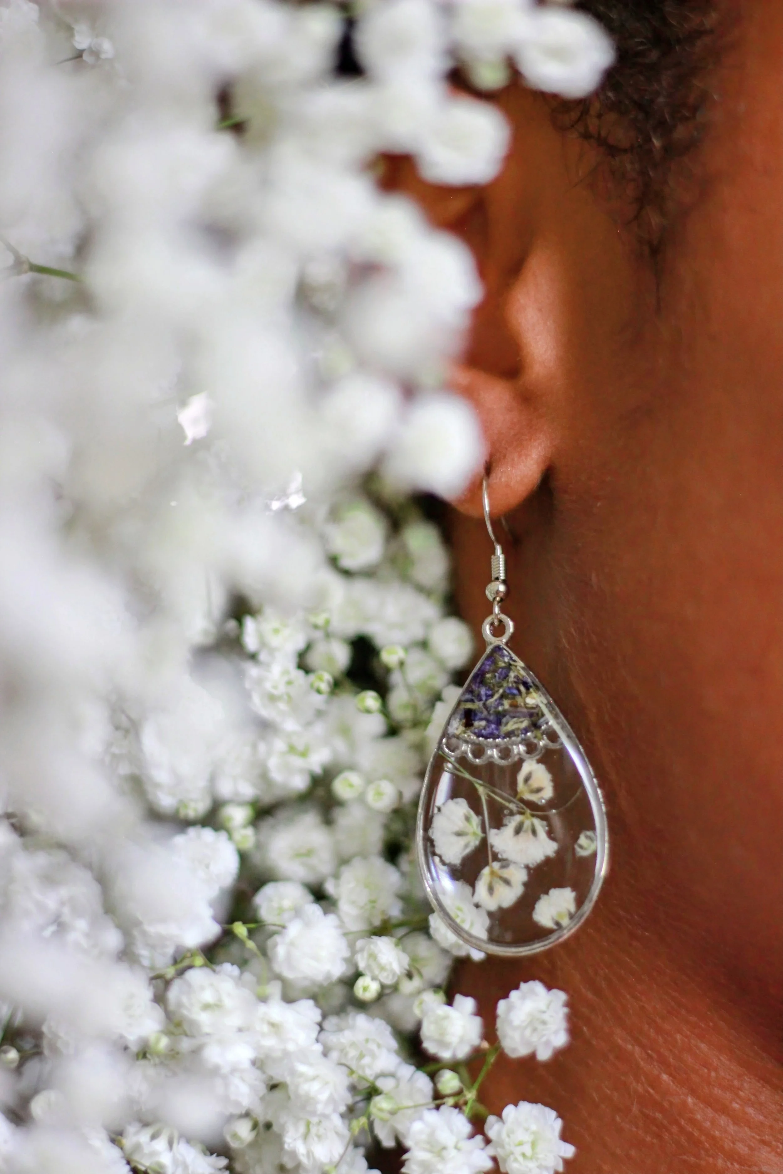 A woman with a brown complexion and curly hair taking a close-up photo of earrings framed by baby's-breath flowers. The person has a silver earring and a teardrop-shaped pendant necklace with white baby's-breath flowers inside.