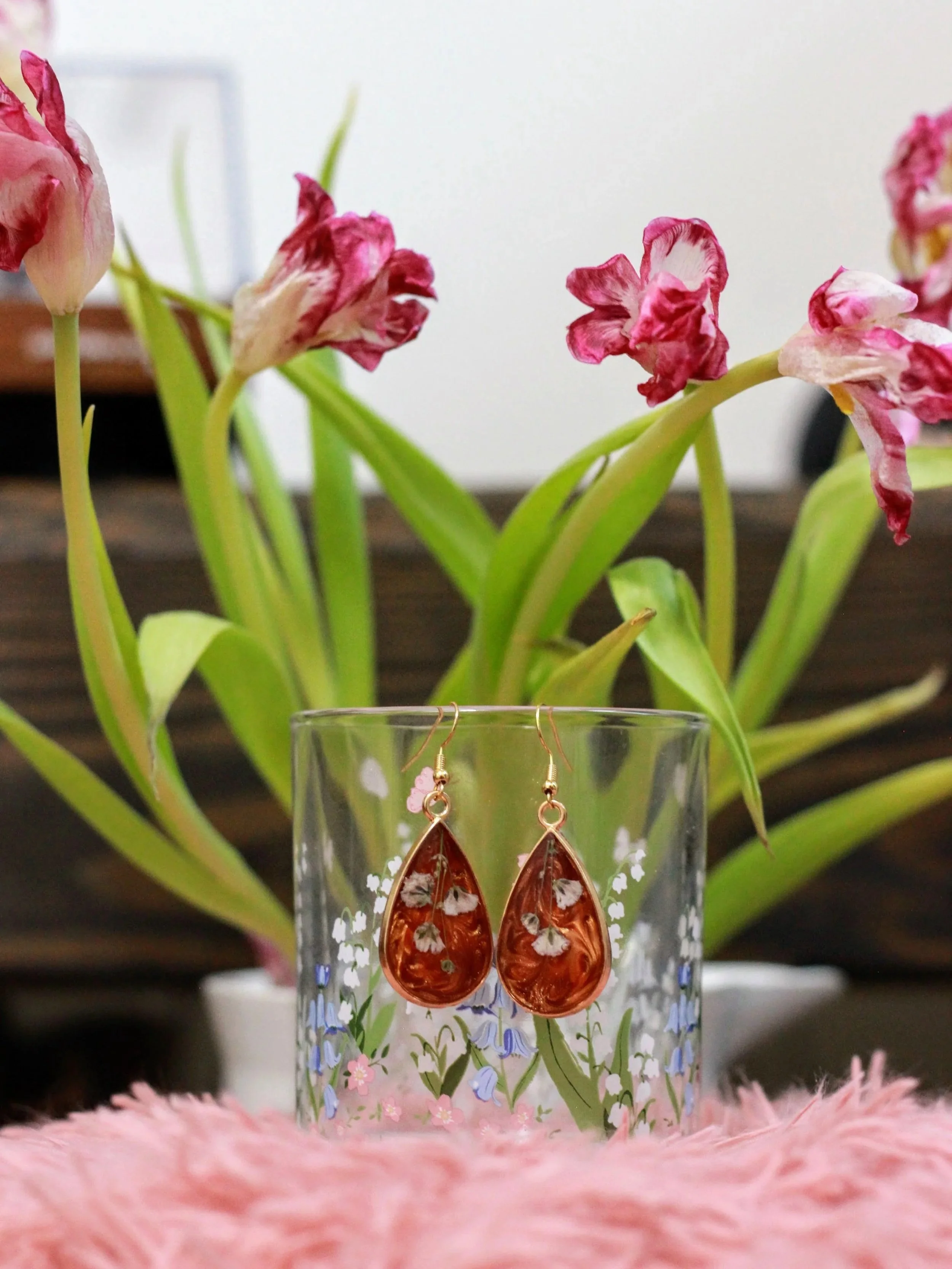 Close-up of a pair of orange teardrop-shaped earrings with white floral patterns hanging from a glass decorated with colorful floral designs. In the background, there are pink and white wilted tulip flowers with green stems and leaves, on a wooden surface with a blurred white wall.