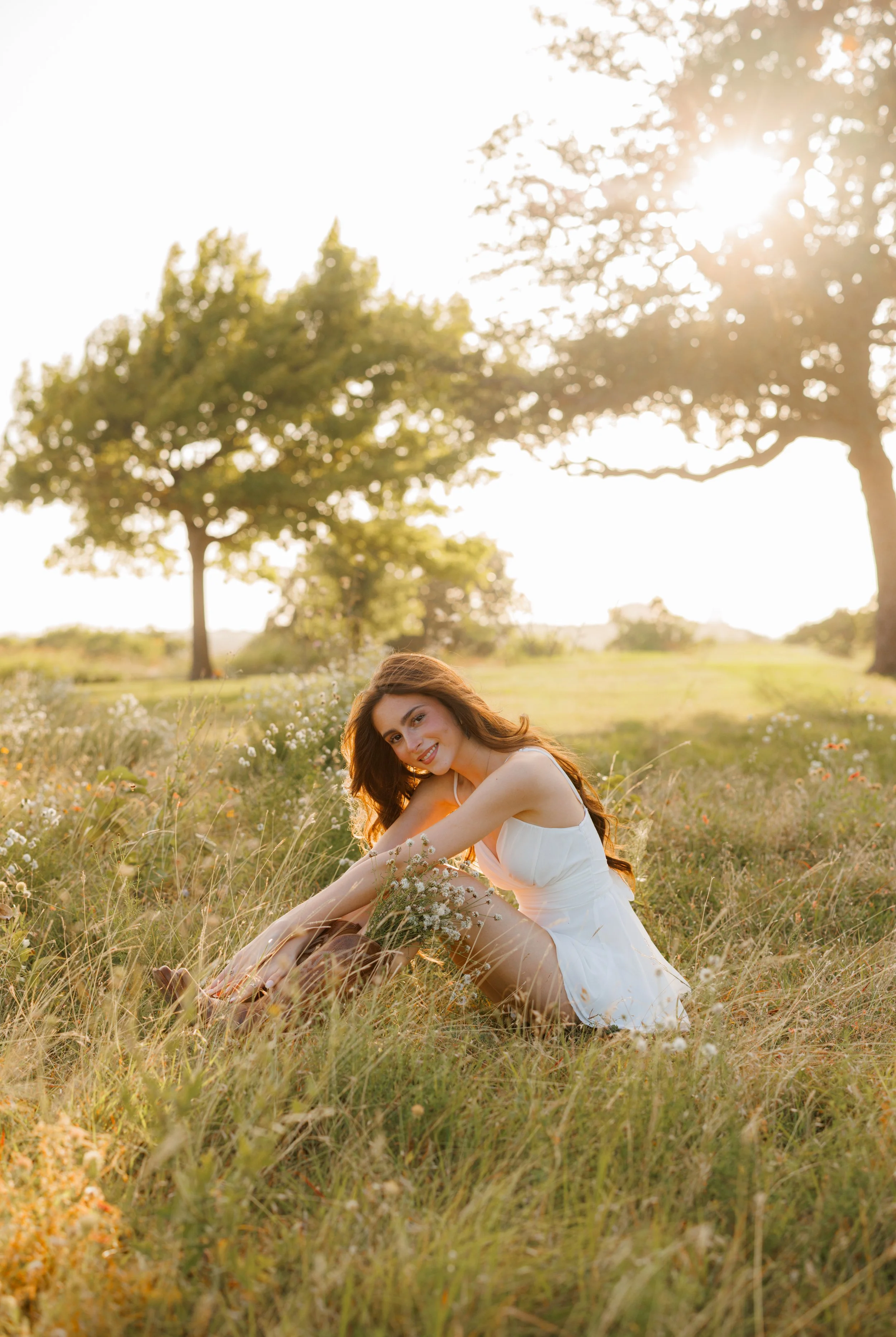 Young woman with long brown hair wearing a white dress, sitting in a meadow of tall grass and wildflowers during sunset with trees in the background.