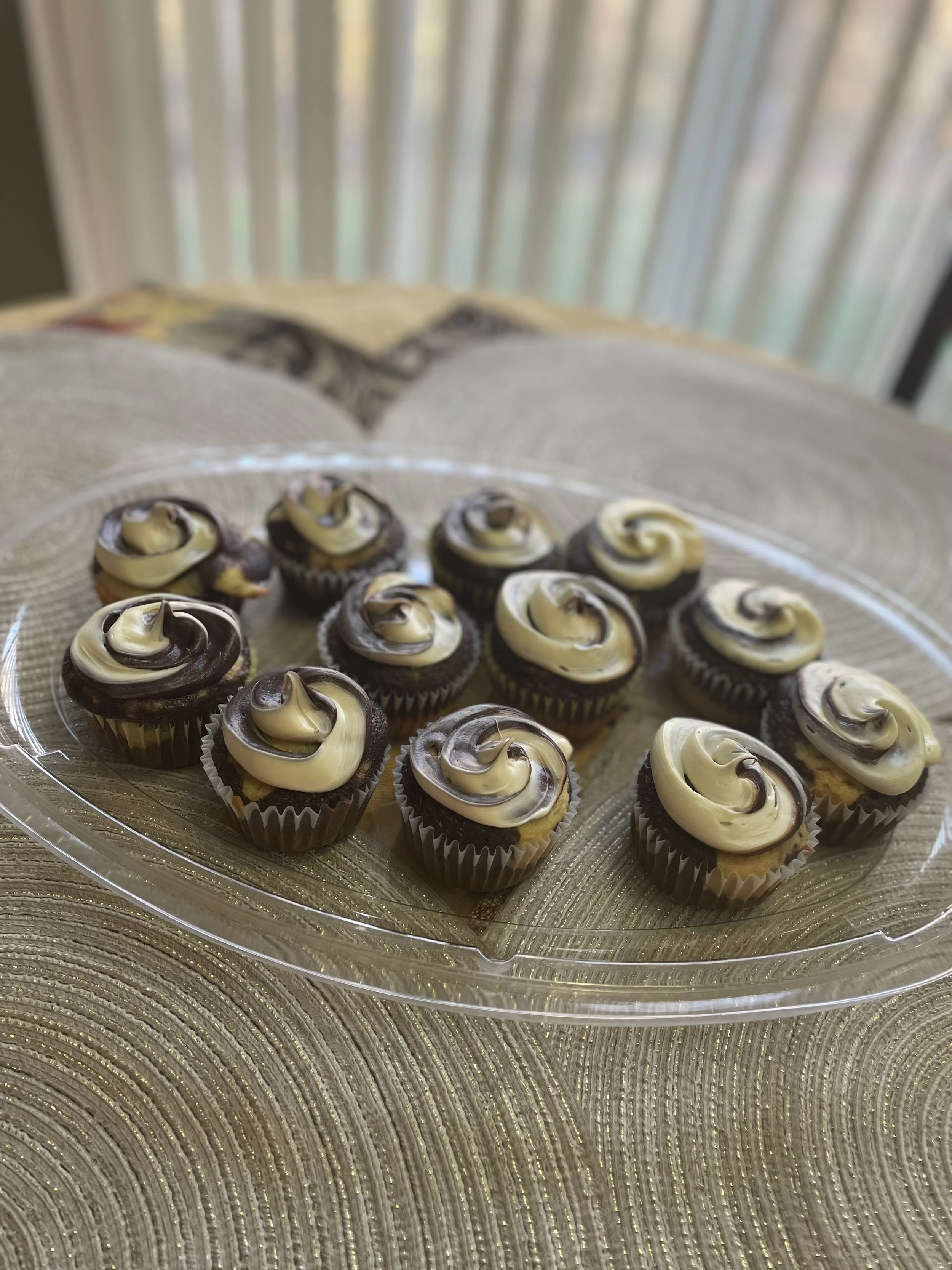 A clear plastic tray of small cupcakes with black and white swirl frosting on top, placed on a textured gold-colored surface.