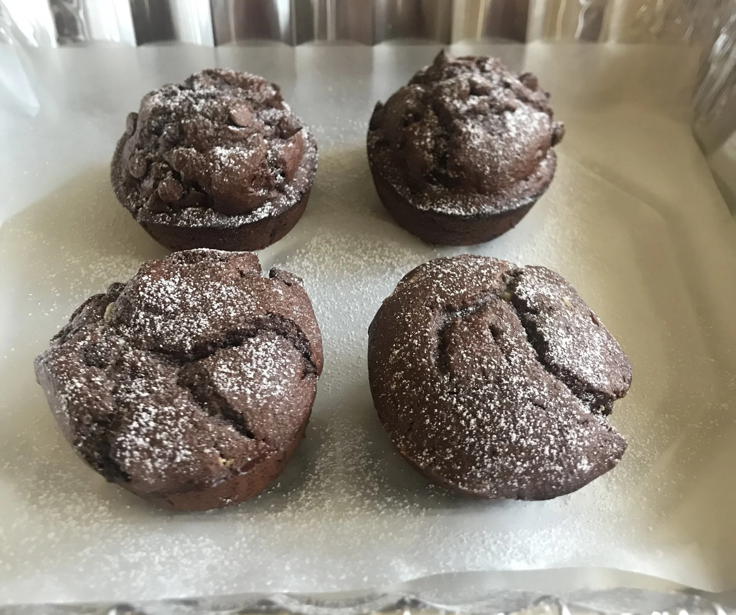 Chocolate muffins dusted with powdered sugar, placed on a baking tray.