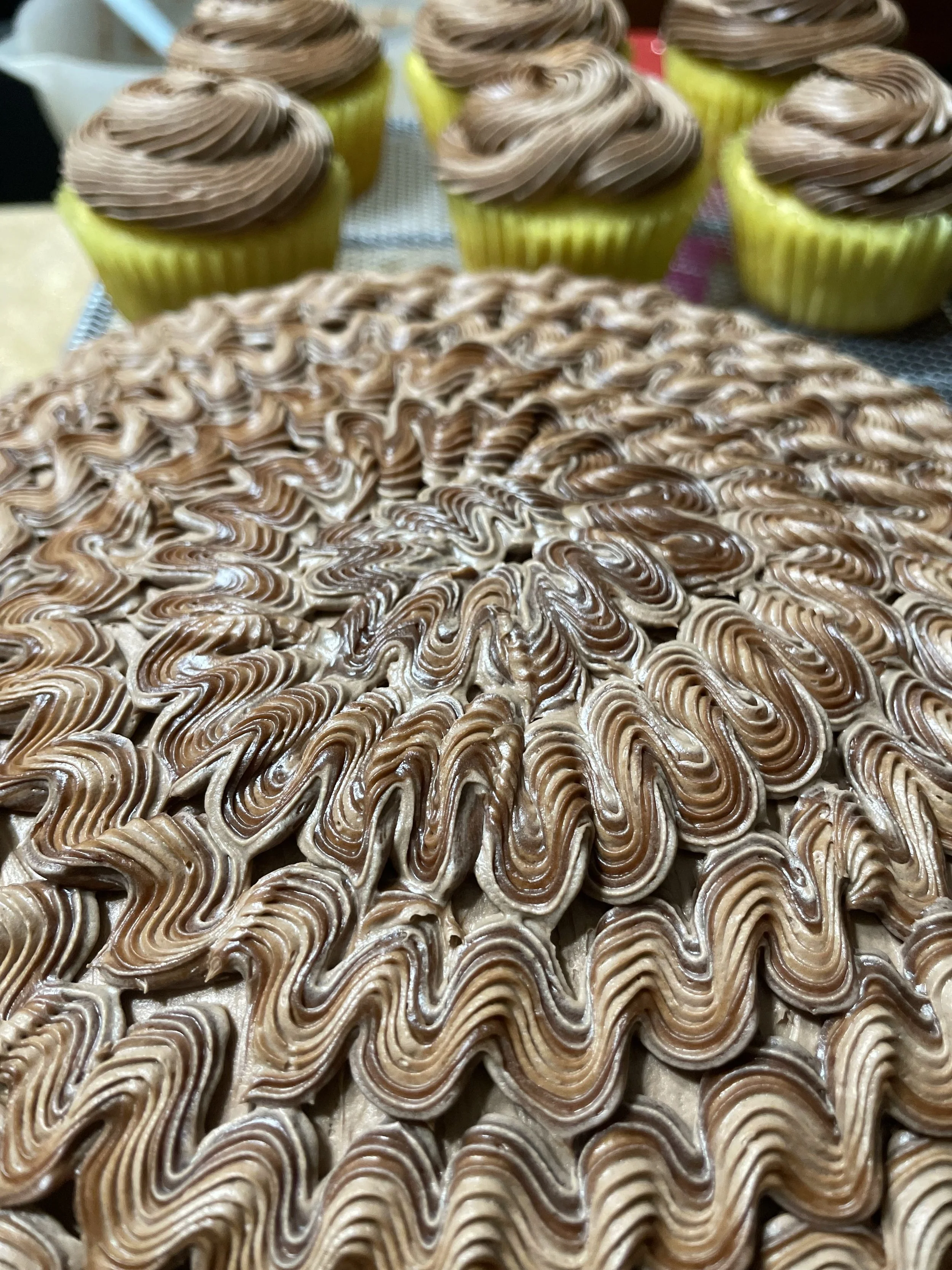 Close-up of a large tray of decorative swirl cookies with chocolate and vanilla frosting, and a row of cupcakes with chocolate frosting on top.