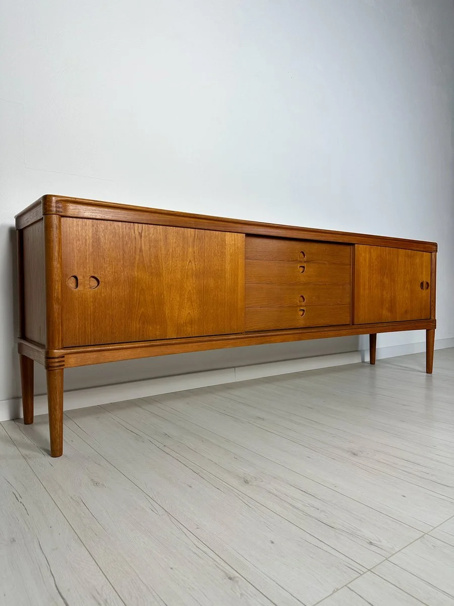 A mid-century modern wooden sideboard or console table with sliding doors and drawers, set against a plain white wall on a light-colored wood floor.