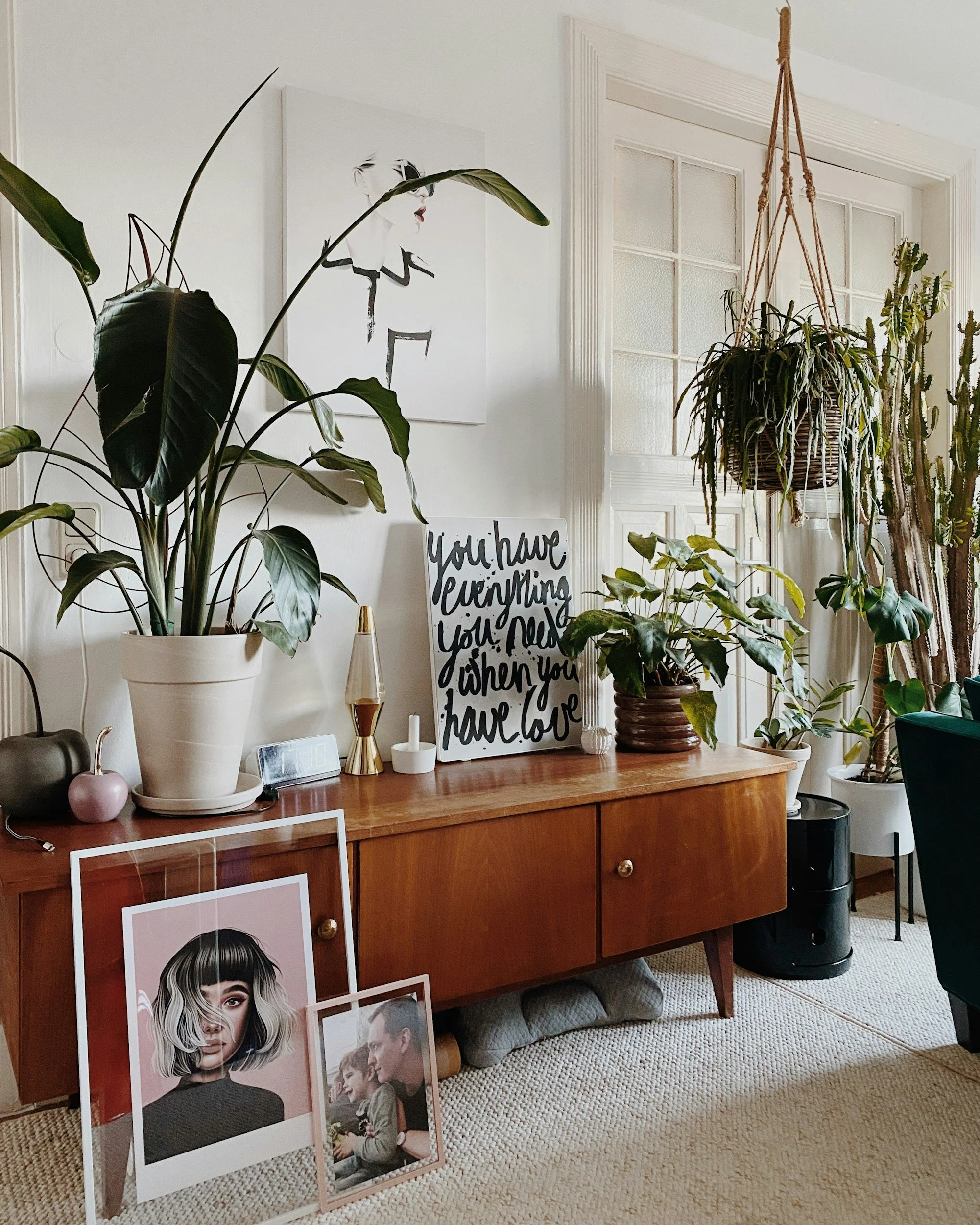 Interior view of a living space with a wooden sideboard decorated with framed artwork, plants, and decorative objects, with additional artwork leaning against the wall on the floor, and a window with white trim in the background.