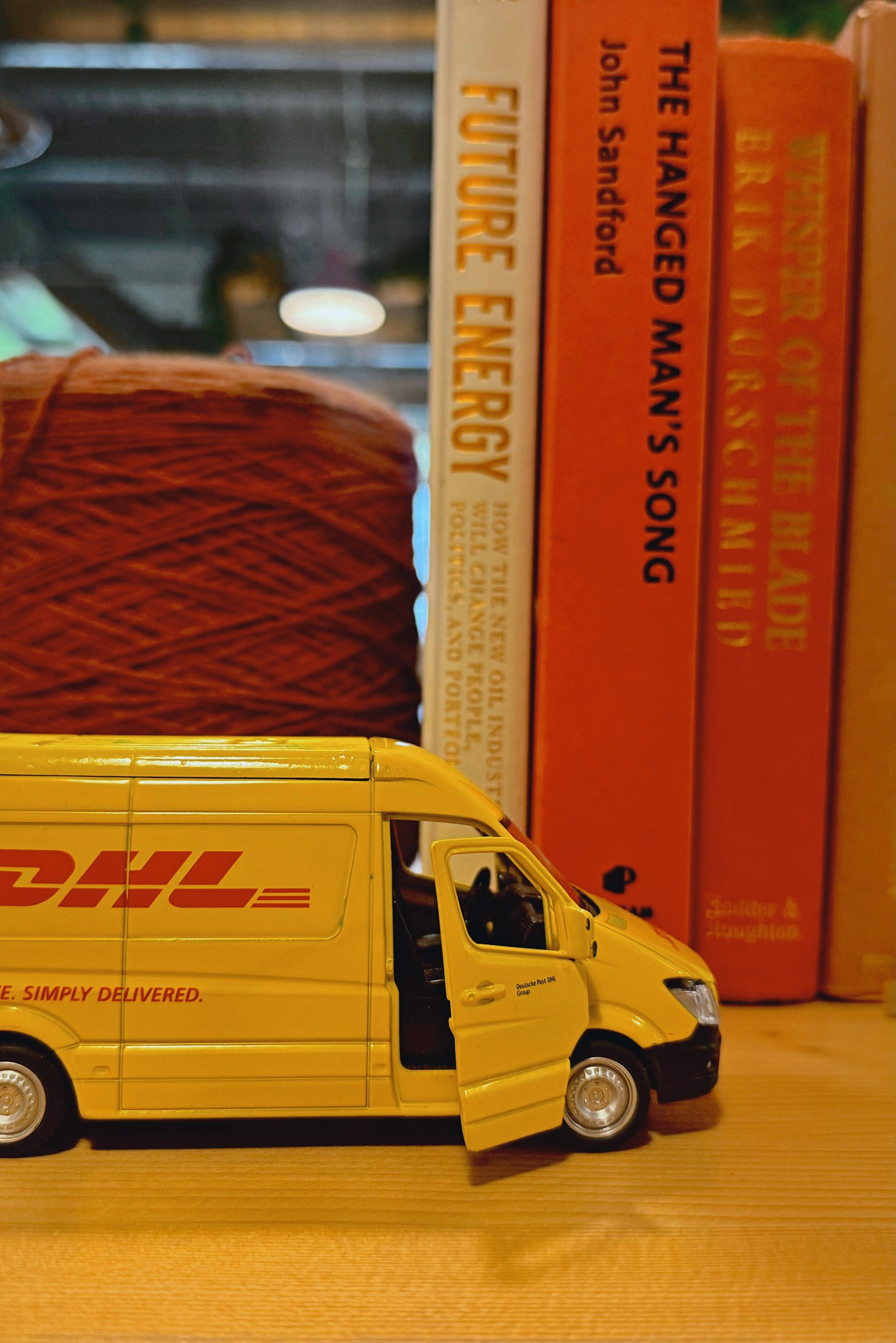A yellow DHL delivery van parked on a wooden surface with a house in the background and books upright behind it.