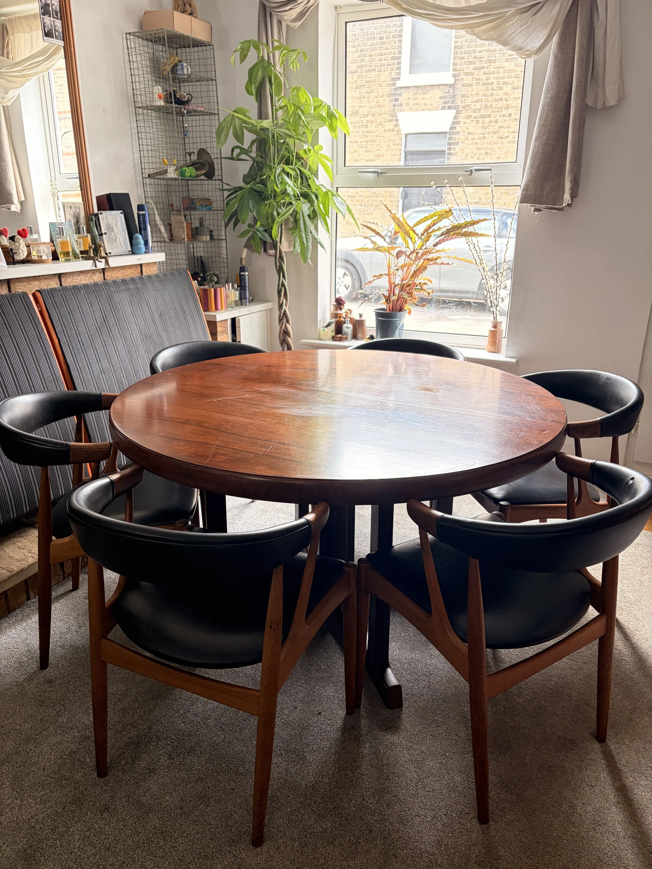 A round wooden dining table surrounded by black and wooden chairs in a cozy room with a large window, indoor plants, and a shelving unit decorated with various small items and books.