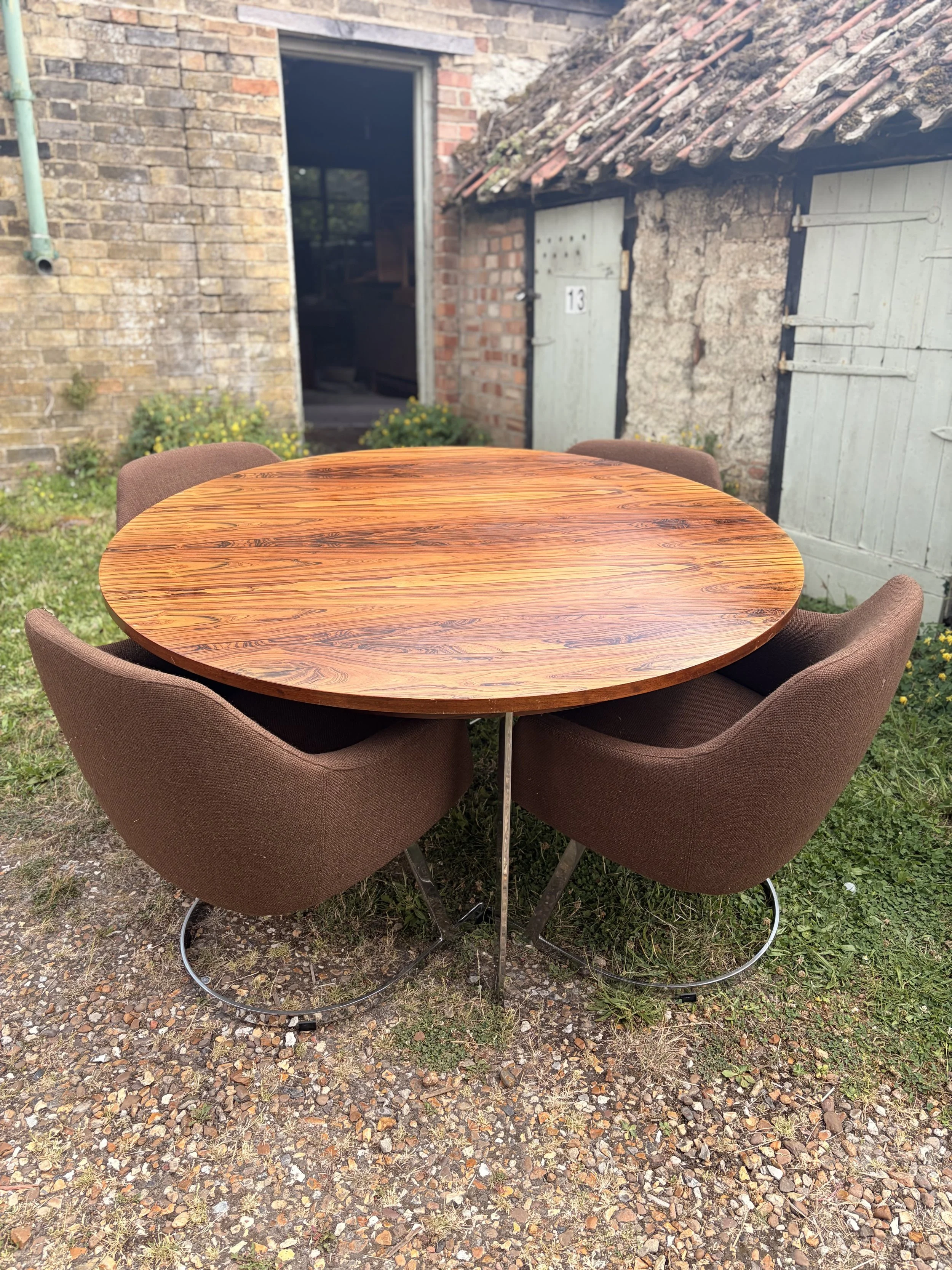 Round wooden table with four brown upholstered chairs on a gravel outdoor surface, with a brick building and a small shed with weathered doors in the background.