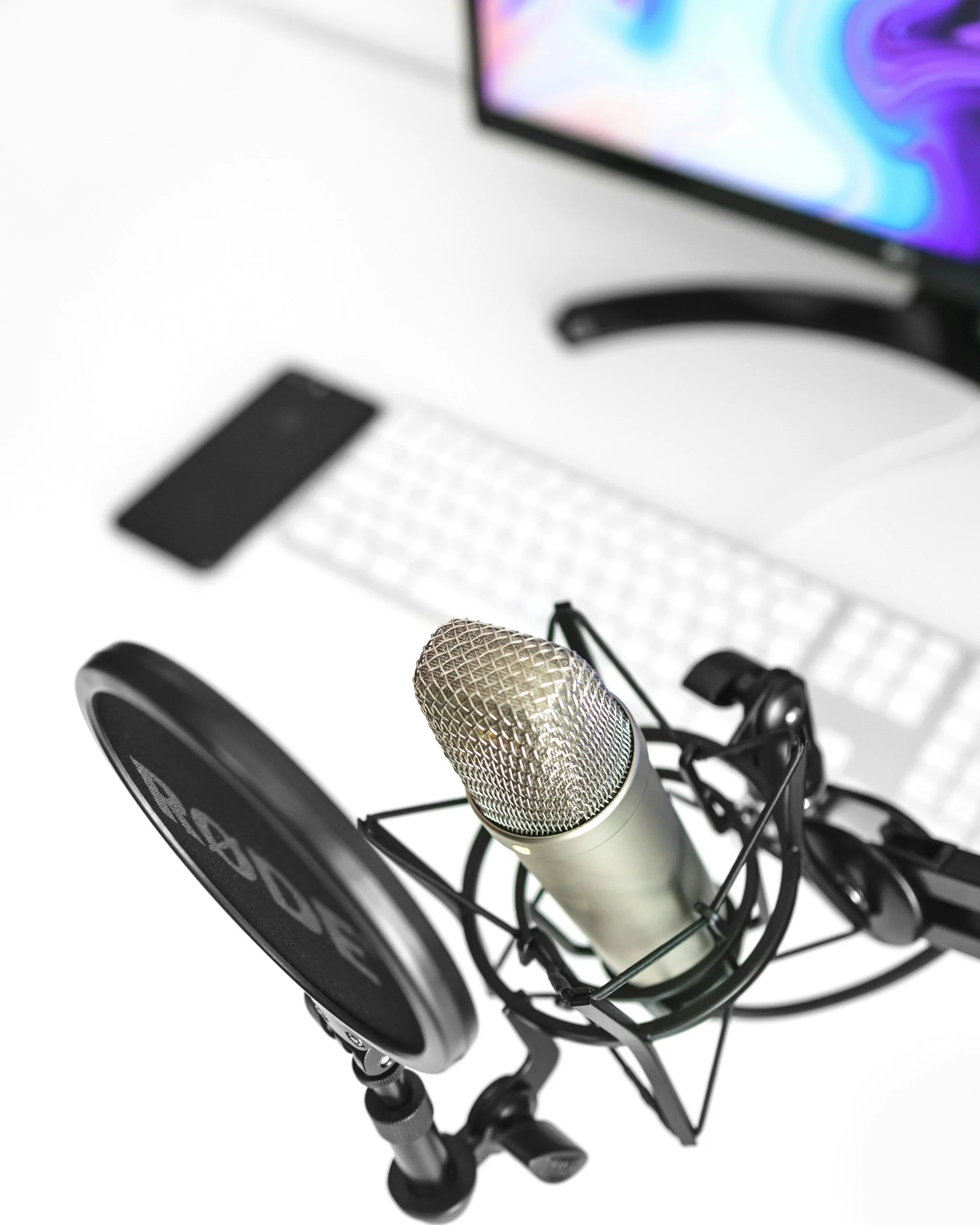 Close-up of a microphone in front of a computer or podcasting setup with a monitor and keyboard in the background.