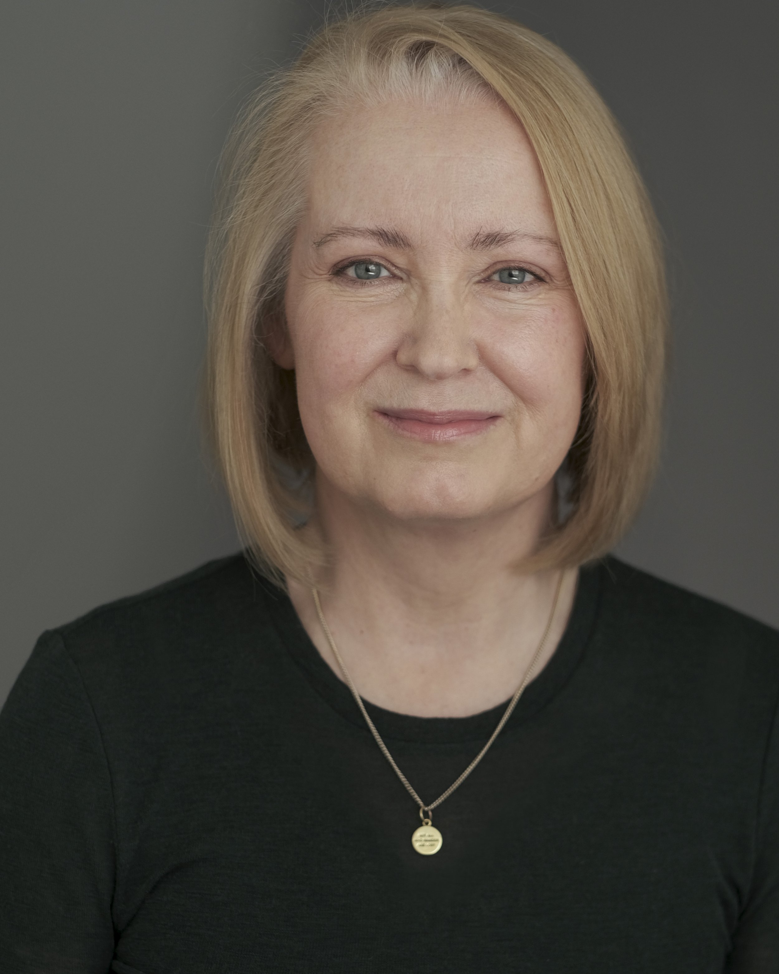 Close-up of a woman with blonde hair, wearing a black top and a silver necklace with a round pendant.
