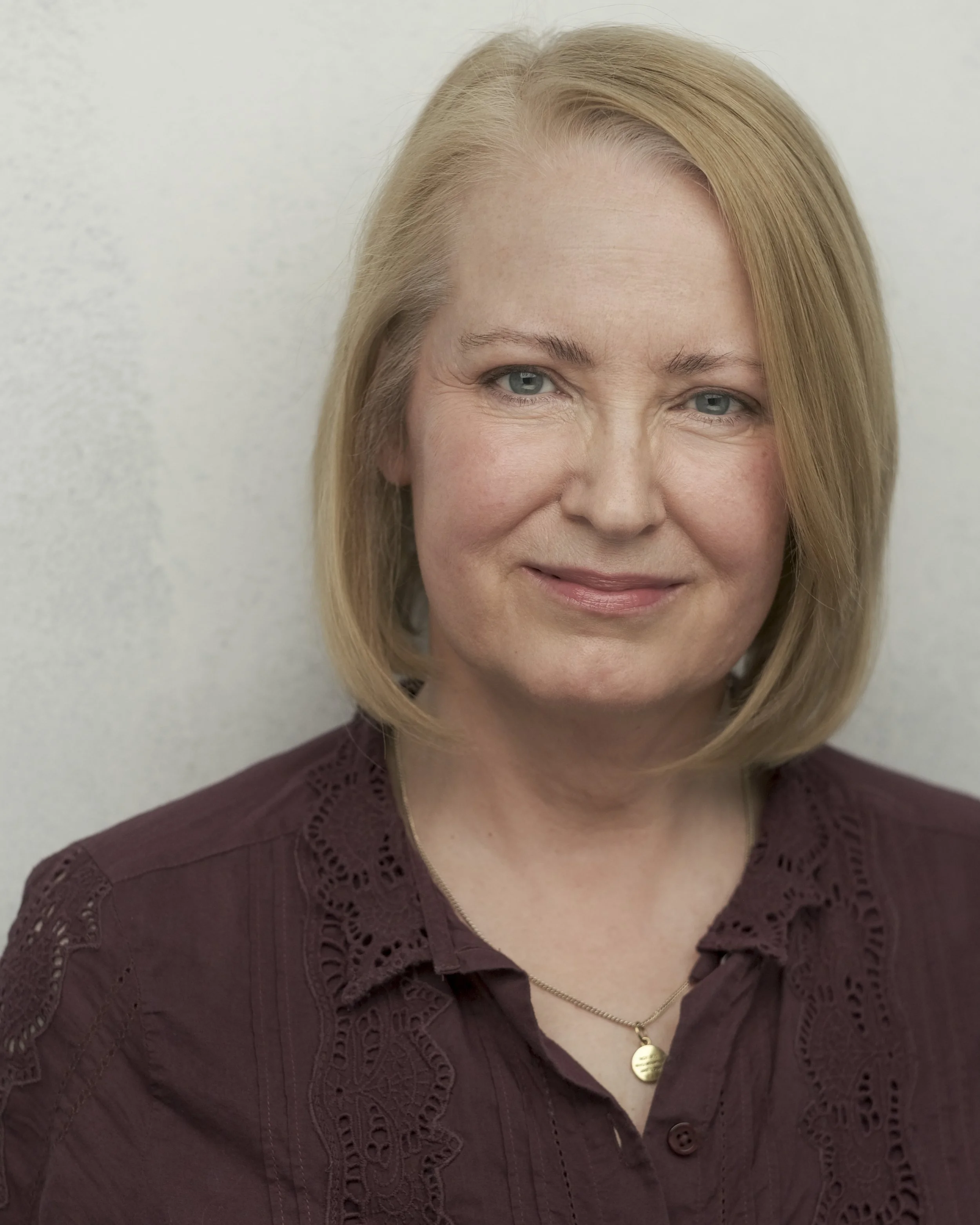 Close-up of a middle-aged woman's face with blonde hair, blue eyes, and a warm smile, wearing a maroon blouse with lace detailing and a gold heart necklace.