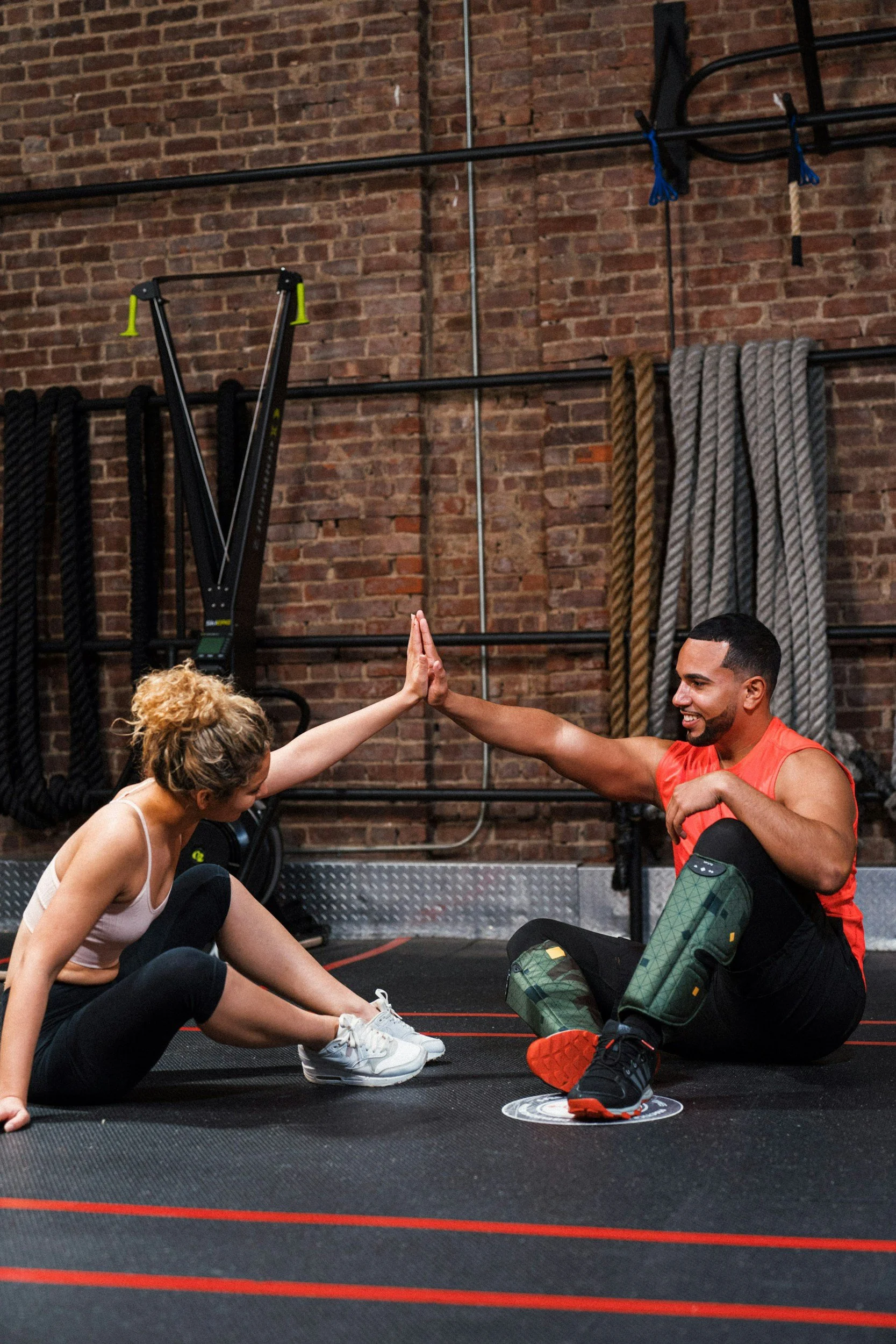 A woman and a man are performing a high-five after a workout in a gym with a brick wall, fitness equipment, and gym ropes in the background.
