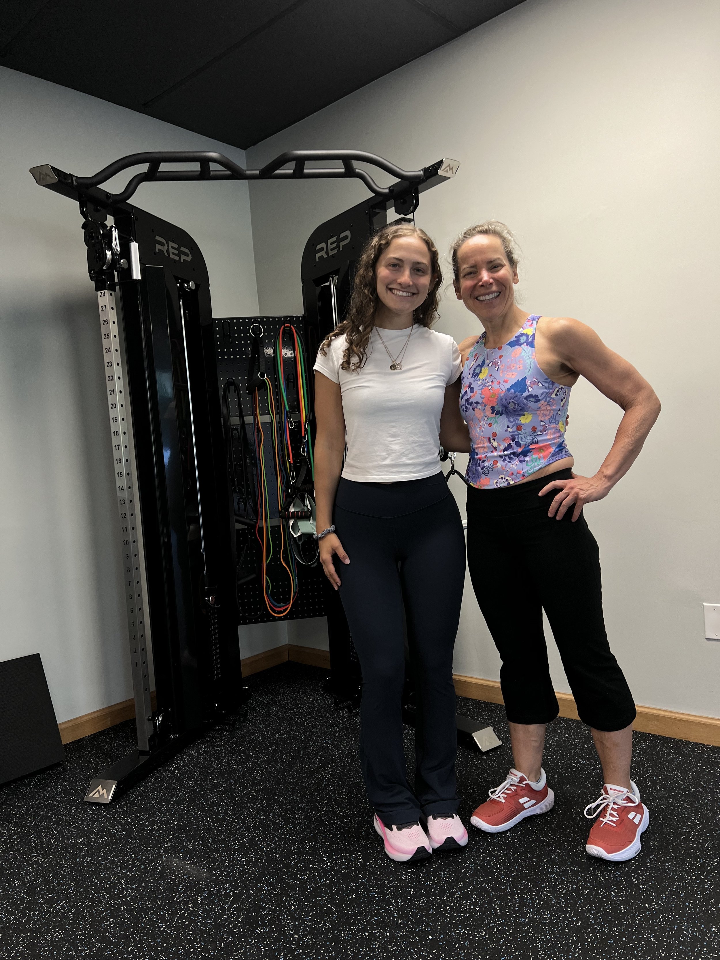 Two women standing in a gym beside strength training equipment, smiling at the camera. One woman is wearing a white t-shirt and dark pants, the other is in a colorful athletic top and black pants. The gym has a black and white speckled floor.