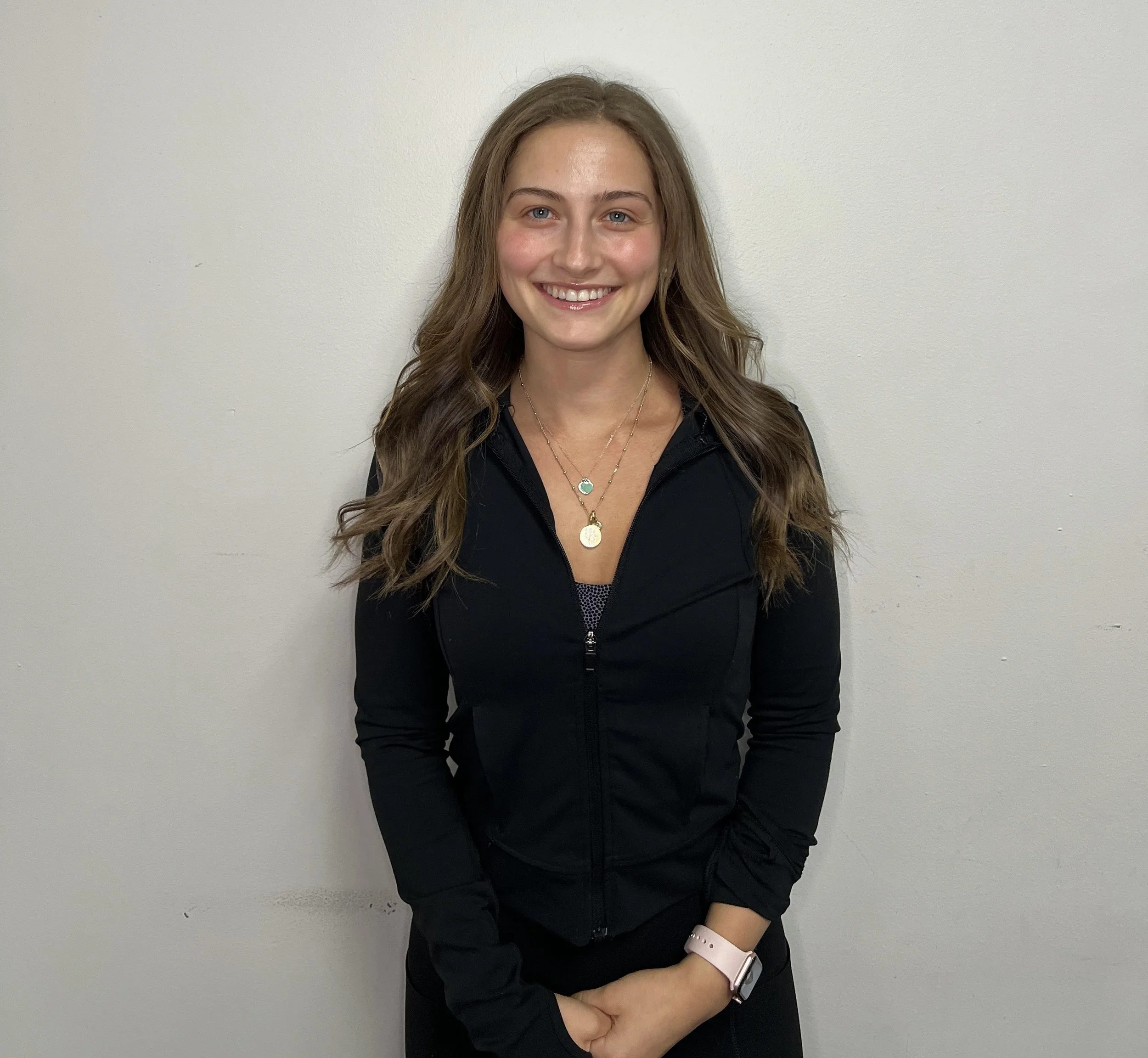 A young woman with long brown hair and blue eyes smiling, wearing a black jacket, gold necklaces, a white watch, and standing against a plain off-white wall.