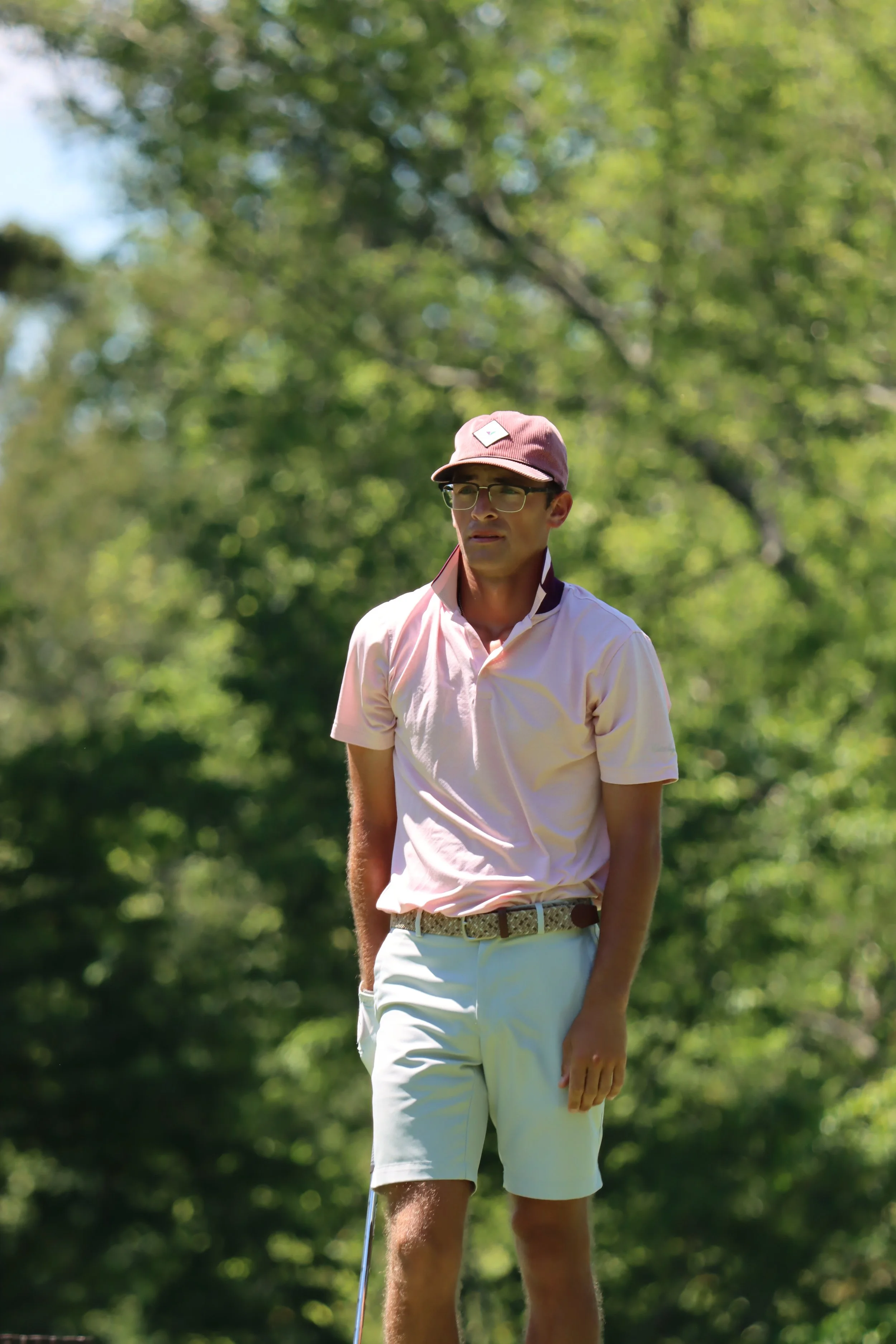 A young man dressed in golf attire, wearing a pink cap, glasses, a light pink polo shirt, and white shorts, standing outdoors on a sunny day with green trees in the background.