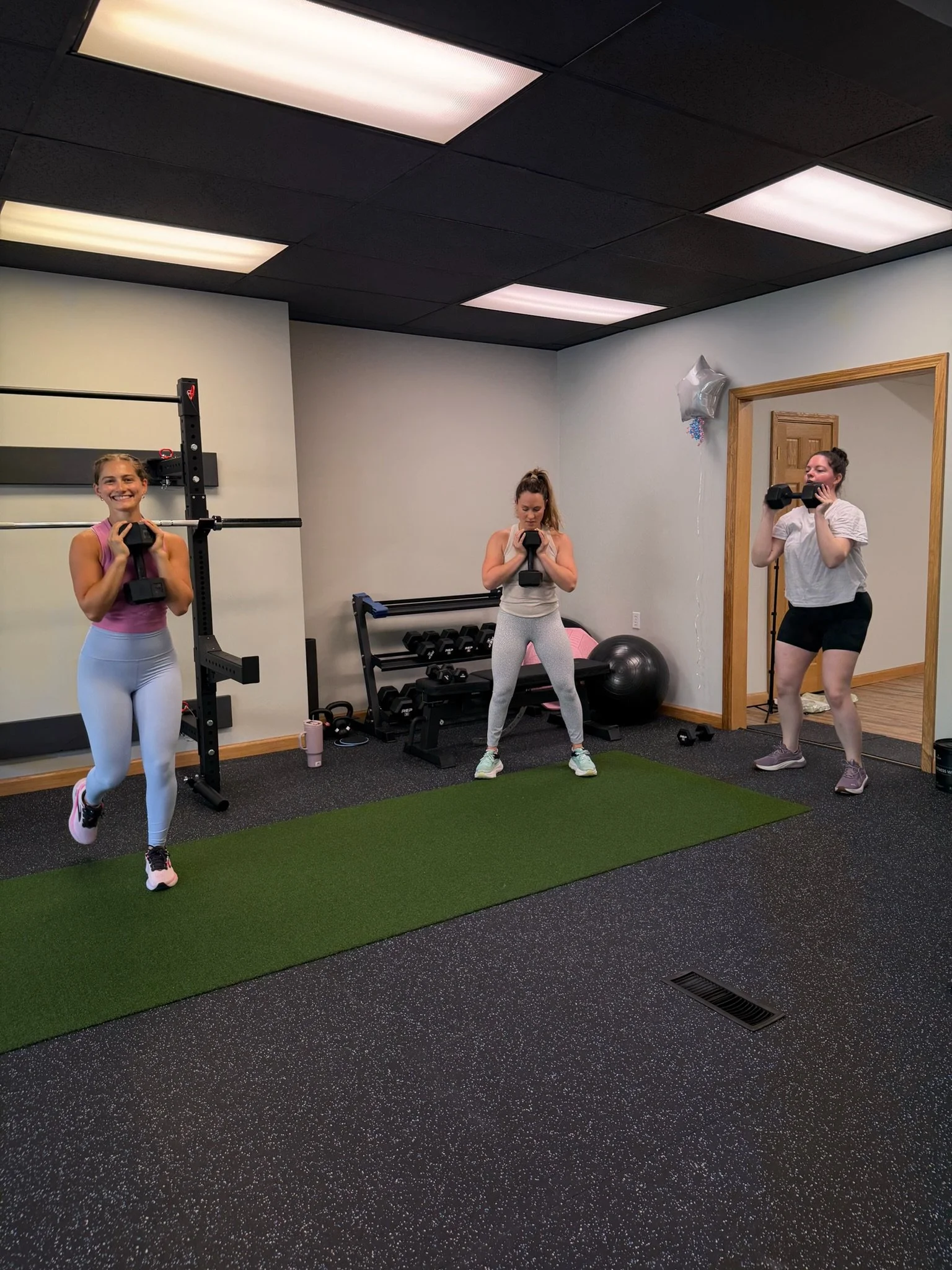 Three women exercising with dumbbells in a gym, standing on a green mat, with gym equipment and a decorative balloon in the background.