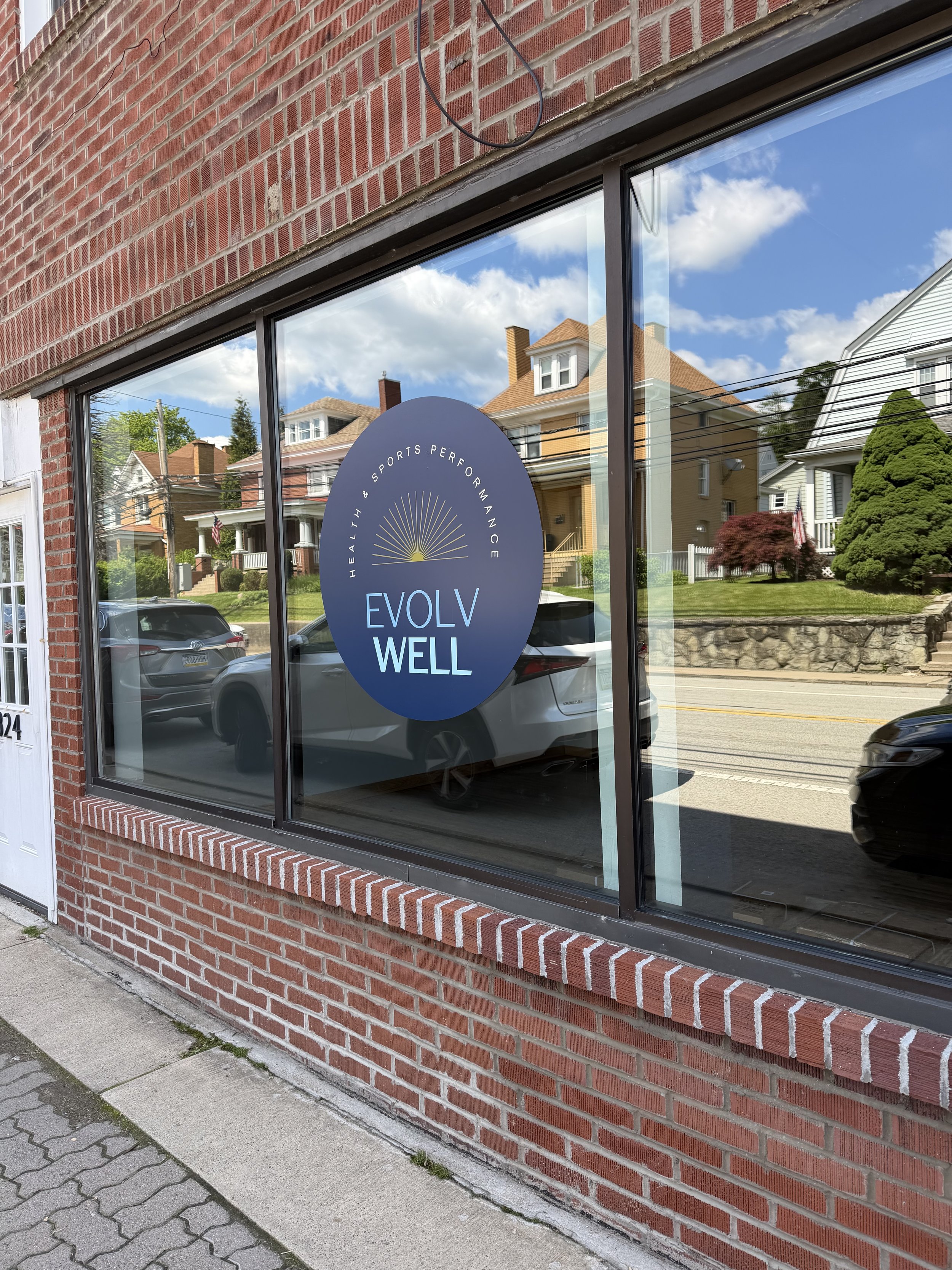The image shows the storefront of a health and sports performance center called 'EVOLV WELL'. The window has a blue circular sign with the logo and the words 'HEALTH & SPORTS PERFORMANCE' around a sunburst graphic.