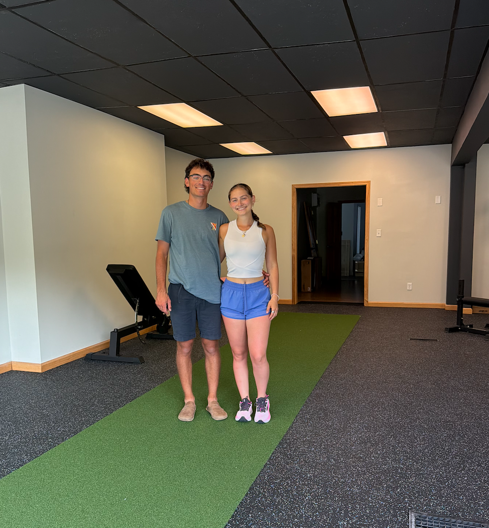 Two people, a young man and a young woman, standing on a green indoor turf, smiling at the camera in a room with black ceiling tiles and gray walls, with exercise equipment visible in the background.