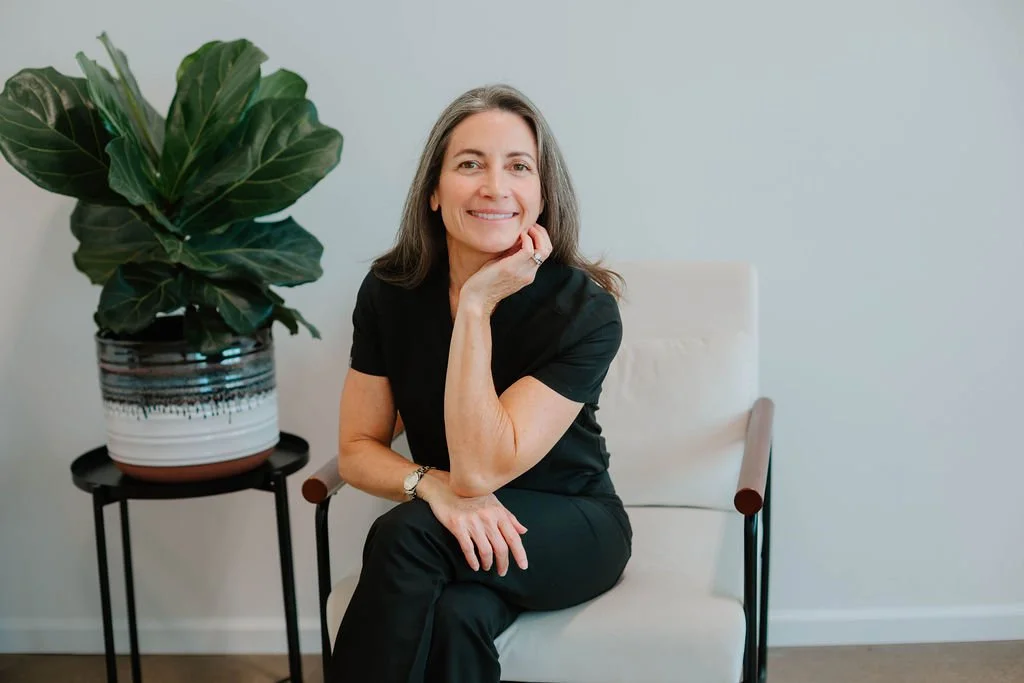 A woman with shoulder-length gray hair sitting on a white chair, smiling and resting her chin on her hand, next to a large potted plant with big green leaves.