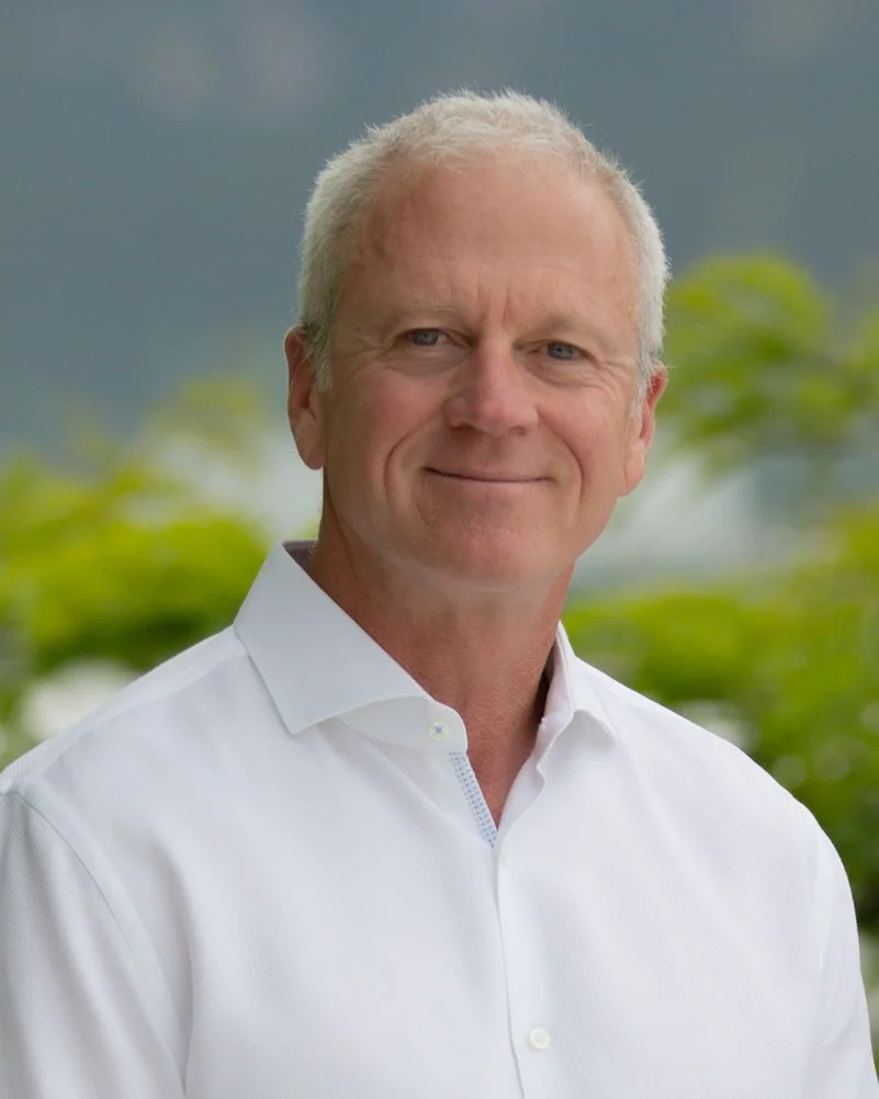 A middle-aged man with short, gray hair, wearing a white dress shirt, standing outdoors with green foliage and cloudy sky in the background.