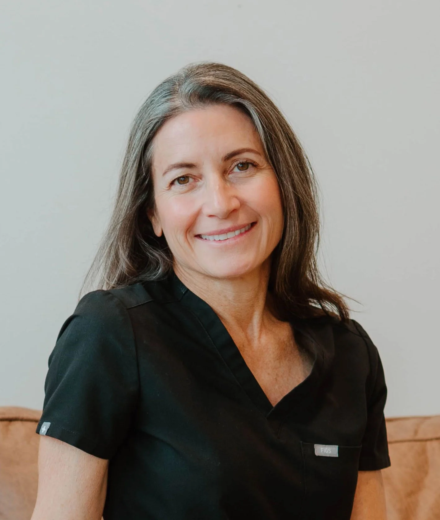A woman with shoulder-length gray and brown hair smiling, wearing a black shirt with a small logo on the sleeve, sitting on a wooden bench in front of a plain light-colored wall.