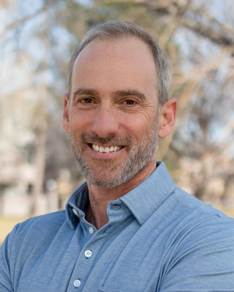 A smiling middle-aged man with short gray hair and a beard, wearing a blue collared shirt, standing outdoors with blurred trees in the background.