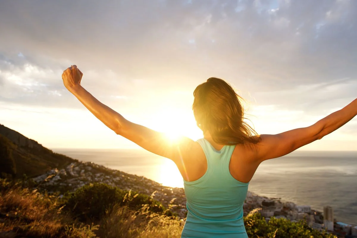 Person standing on a hill with arms raised, overlooking the ocean at sunset.