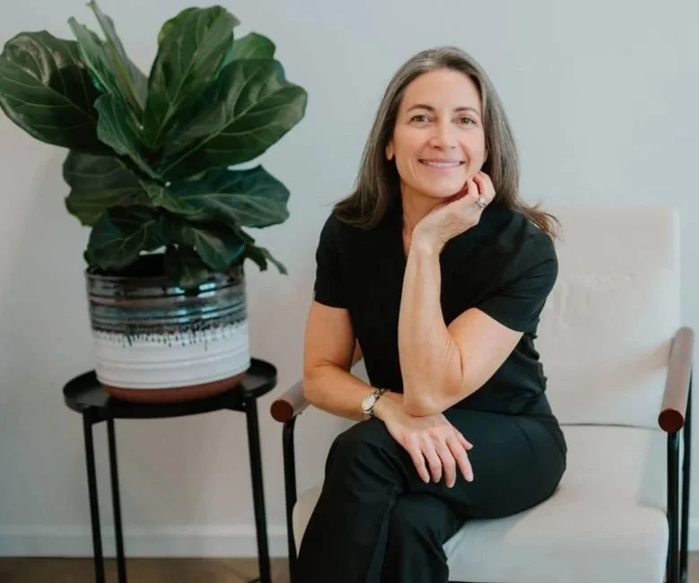 A smiling woman with gray hair, wearing a black outfit, sitting on a white armchair next to a potted plant with large green leaves.