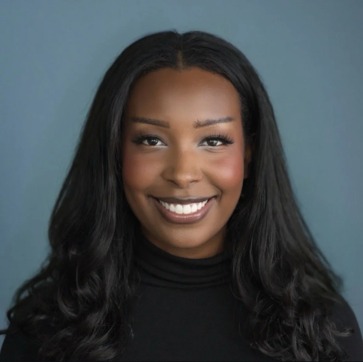 A young African American woman with long black wavy hair, wearing a black turtleneck, smiling against a blue background.