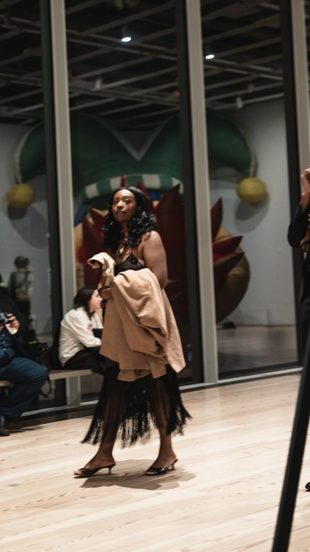 A woman in a black dress and clear heels is standing on a wooden floor, holding a beige coat, with people sitting behind her and colorful artwork reflected in glass behind her.