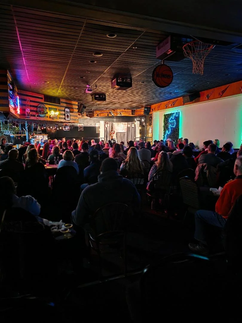 Comedy show in a dimly lit bar or club with a stage, audience seated facing the stage, neon signs on walls, and a basketball hoop hanging from the ceiling. Decatur Conference Center & Hotel
