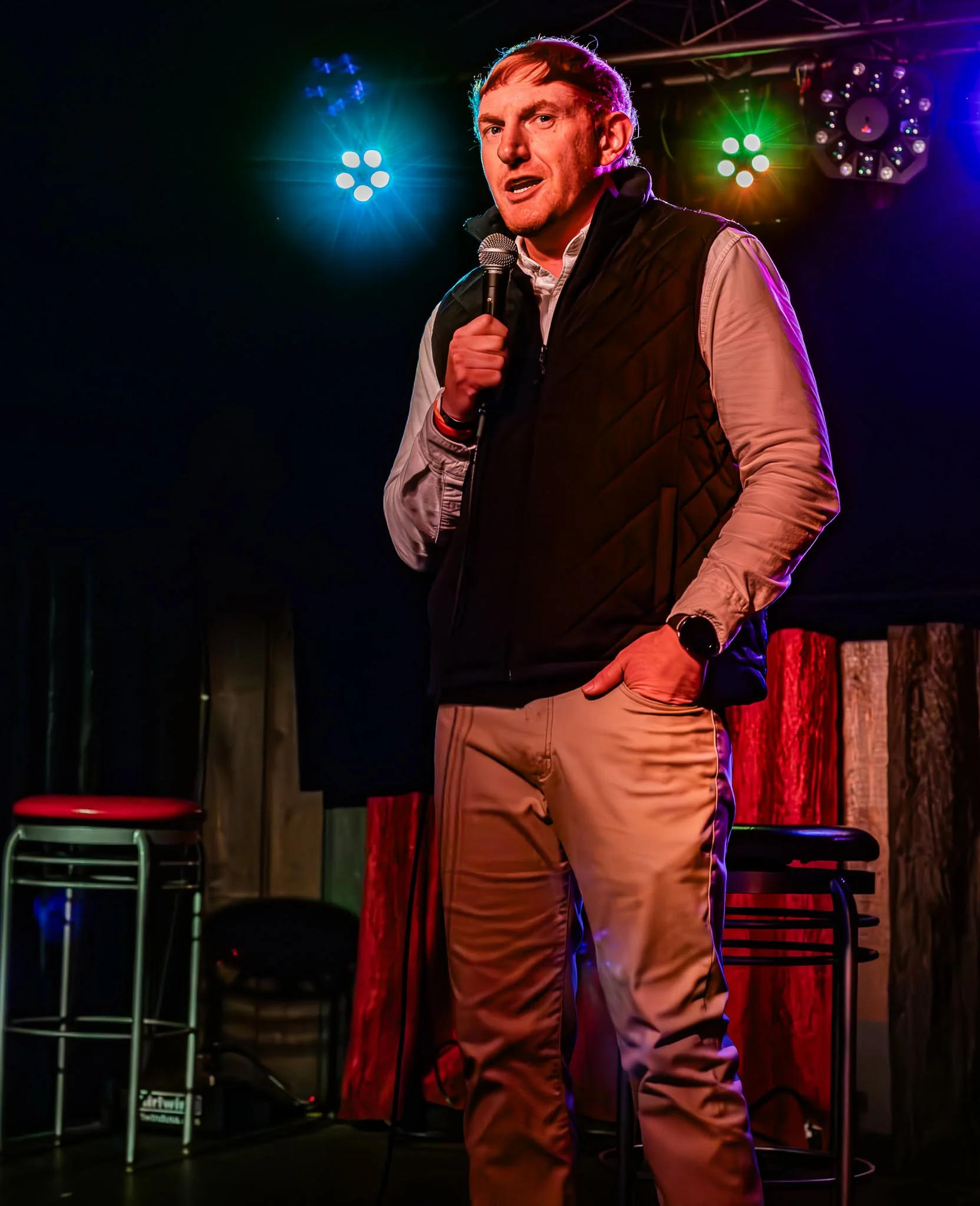 A man holding a microphone, standing on stage with colorful stage lights shining behind him, wearing a black vest and beige pants.