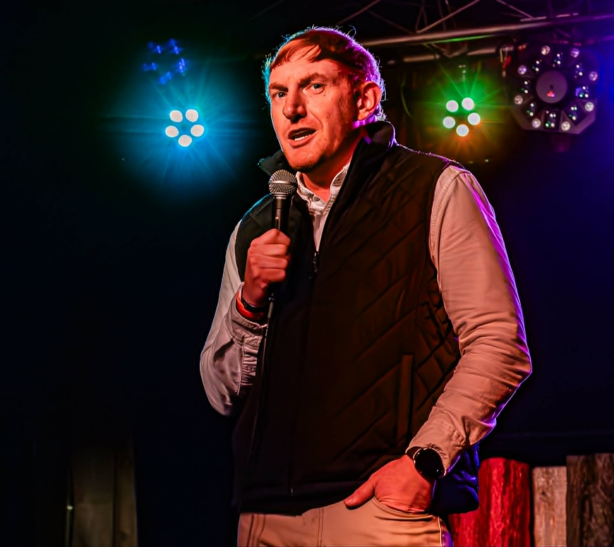 A man holding a microphone on stage with colorful stage lights shining behind him. Craig Antonio is the comedian pictured.
