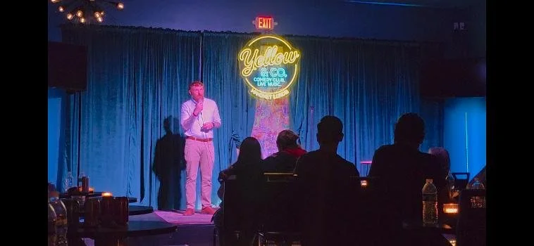 A comedian stands on stage at Yellow Joe's Comedy Club, illuminated by a large neon sign, with an audience seated in front.