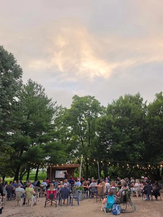 Outdoor movie night scene with people seated on chairs in a park under trees, watching a film on a large screen, with string lights overhead at dusk.