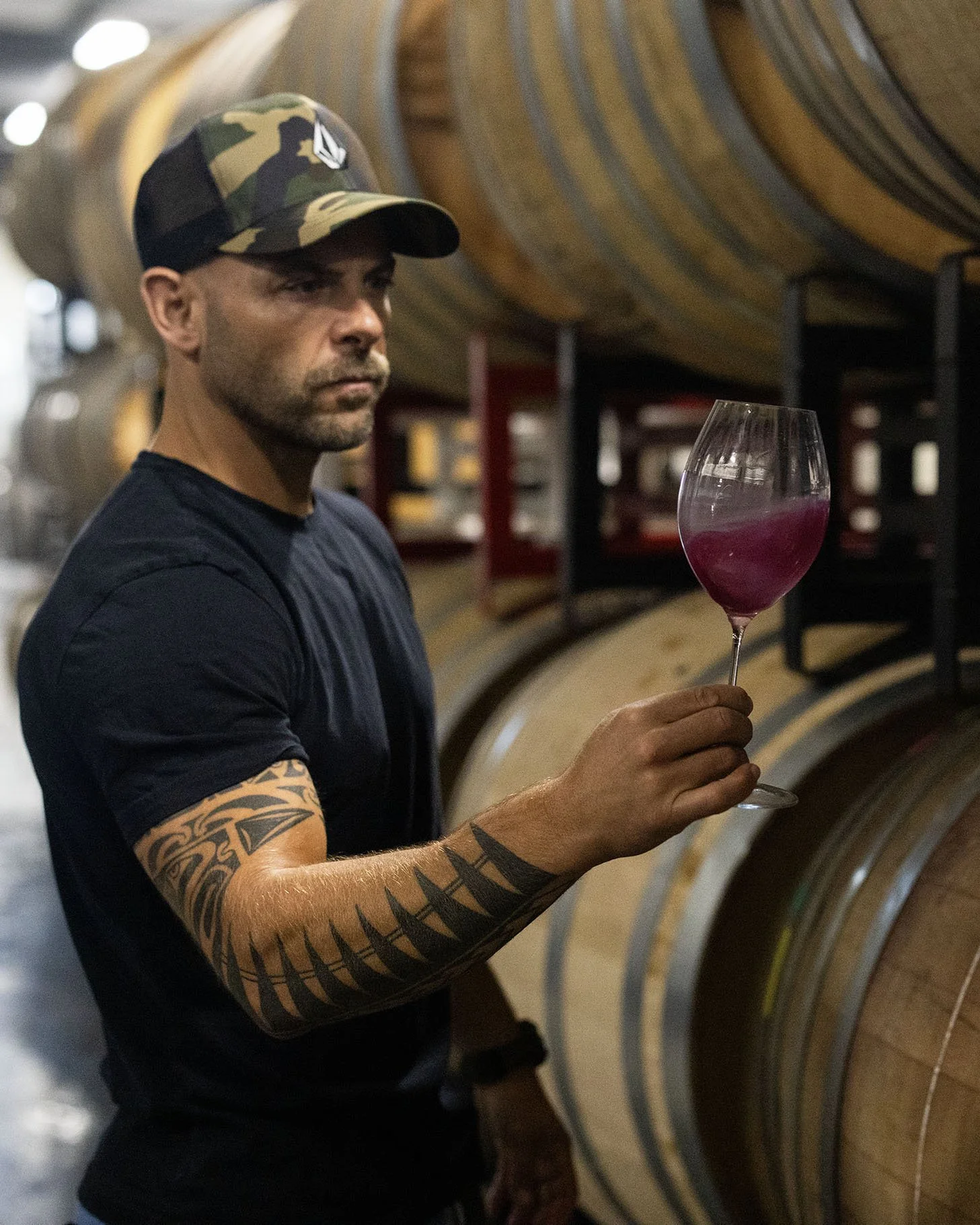 A man wearing a camouflage hat and a black t-shirt holding a wine glass with pinkish-red wine inside, standing in front of large wooden wine barrels in a wine cellar.
