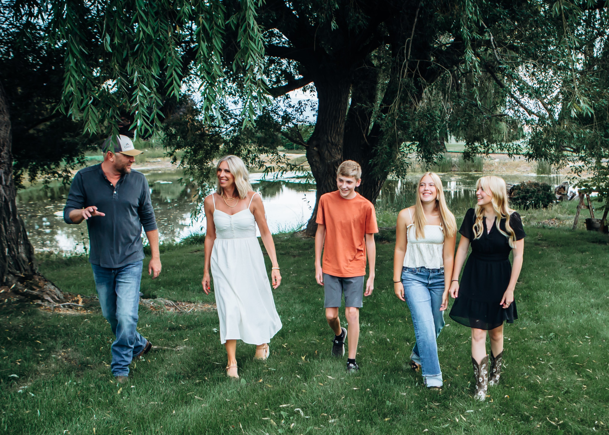 A group of five people walking outdoors near a pond with trees in the background. The group consists of one man and four women and teenagers, all smiling and enjoying a sunny day.