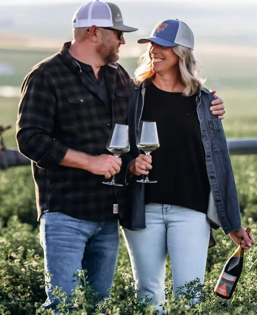 A man and woman stand close together outdoors, holding glasses of wine, smiling and looking at each other, with a woman holding a wine bottle. They are in a vineyard during daytime.