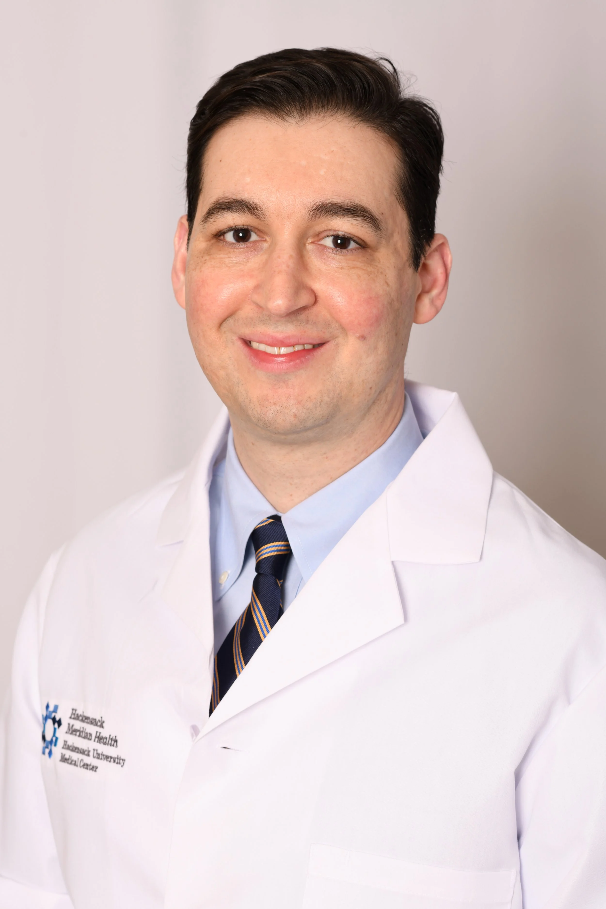 A doctor in a white coat with a logo for Biscayne Medical Center, petite striped tie, light blue shirt, smiling in front of a plain background.