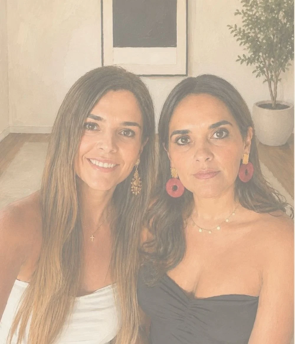 Dos mujeres con cabello largo, una con delgada blusa blanca y la otra con vestido negro, posando juntas en un interior con pared clara, cuadro y planta decorativa visible.