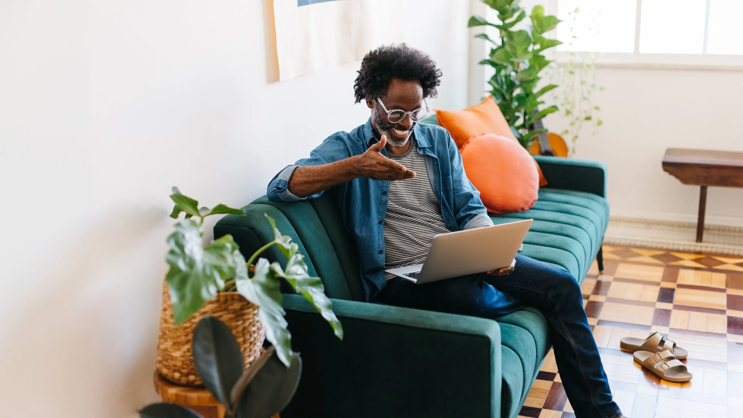 A man with curly hair and glasses sitting on a teal sofa, smiling and gesturing while looking at a laptop in a sunlit room with houseplants, orange pillow, patterned rug, and wooden side table.