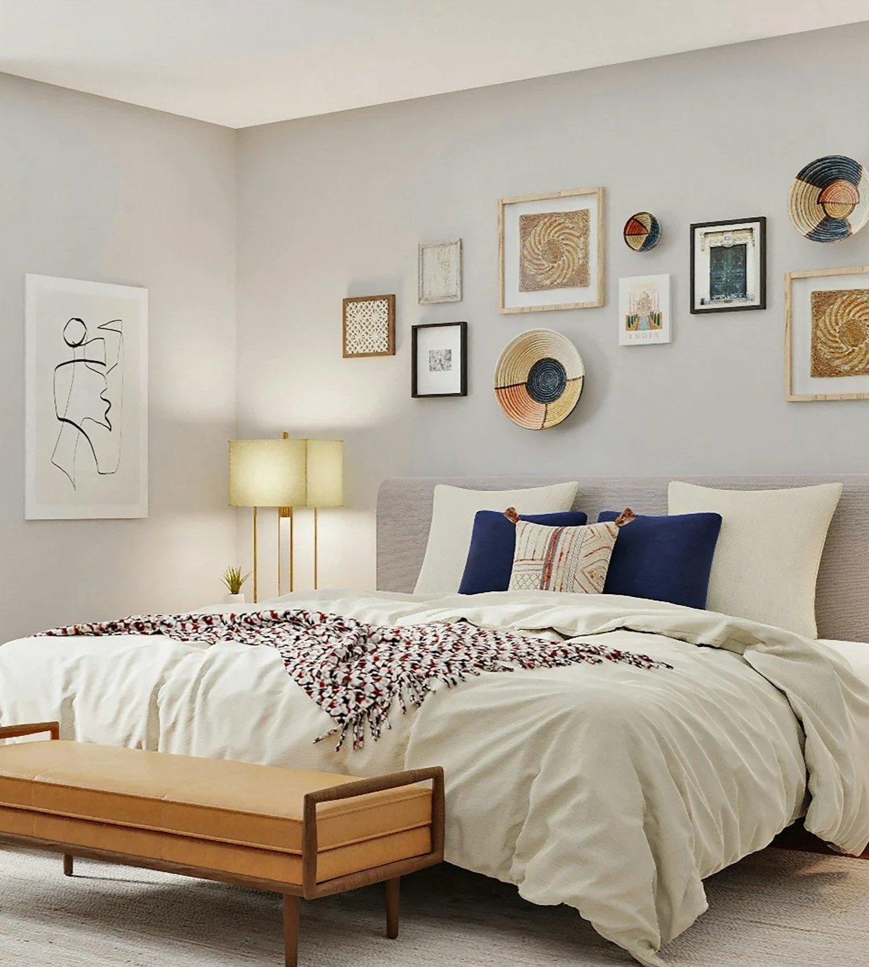 A neatly made bed with white bedding, navy blue and patterned pillows, and a decorative throw blanket. The bed has a padded headboard, and a wooden bench sits at the foot of the bed. Behind the bed, a gallery wall features various framed artwork, including woven baskets, abstract art, and photographs. To the left, a nightstand holds a small potted plant and a double-lamp with a white shade. The room has light gray walls and a beige carpet.